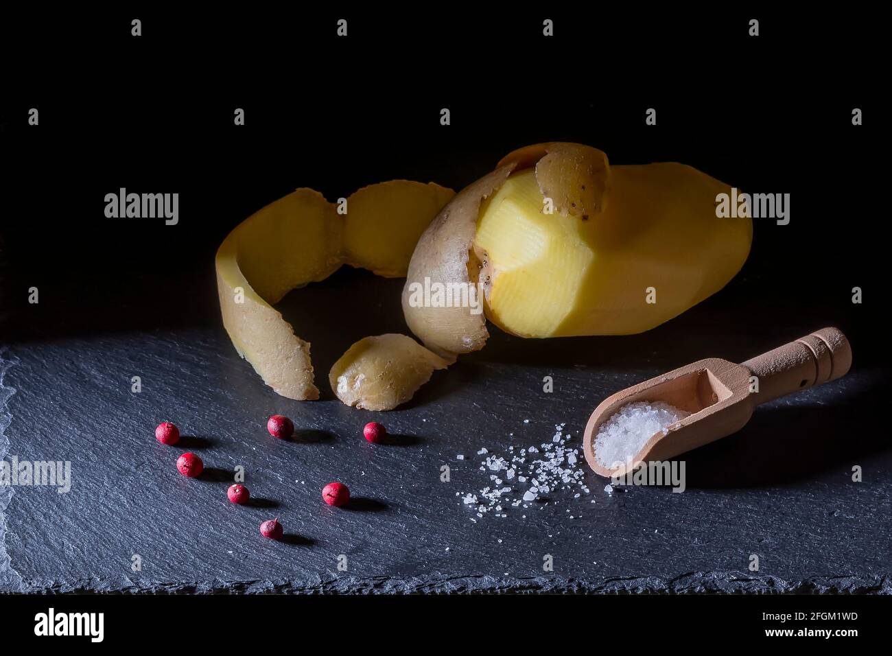 Une pomme de terre pelée avec la peau reposant sur le dessus, des baies rouges et une cuillère en bois pleine de grains de sel sur une plaque d'ardoise Banque D'Images