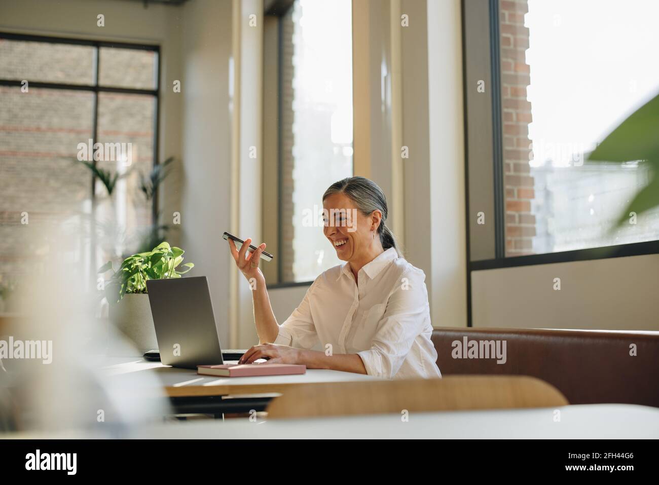 Femme souriante utilisant le téléphone et l'ordinateur portable au bureau. Femme adulte de taille moyenne parlant sur un téléphone à haut-parleur dans un espace de travail de collègue . Banque D'Images Femme souriante utilisant le téléphone et l'ordinateur portable au bureau. Femme adulte de taille moyenne parlant sur un téléphone à haut-parleur dans un espace de travail de collègue . Banque D'Images