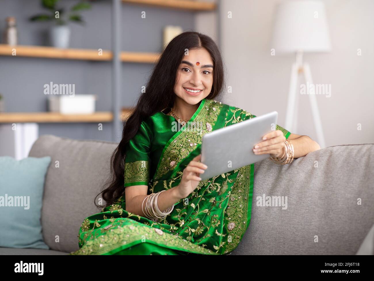 Portrait d'une femme indienne heureuse en robe traditionnelle sari en utilisant tablette sur le canapé à la maison Banque D'Images