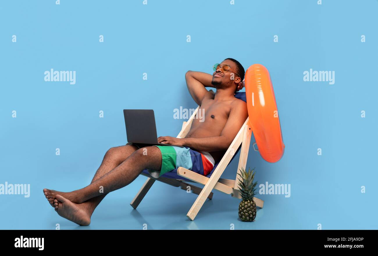 Un homme noir décontracté se détend dans une chaise longue avec ordinateur portable, travaillant en ligne depuis un paradis tropical, fond bleu Banque D'Images