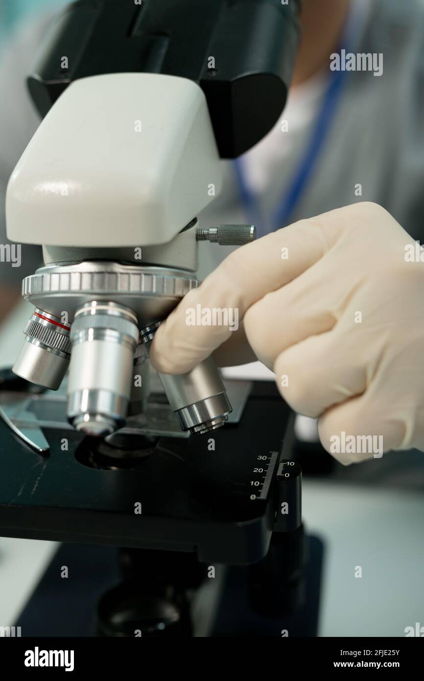 Gros plan d'un scientifique méconnaissable avec des gants qui ajustent la lentille de l'objectif avant recherche au microscope en laboratoire Banque D'Images