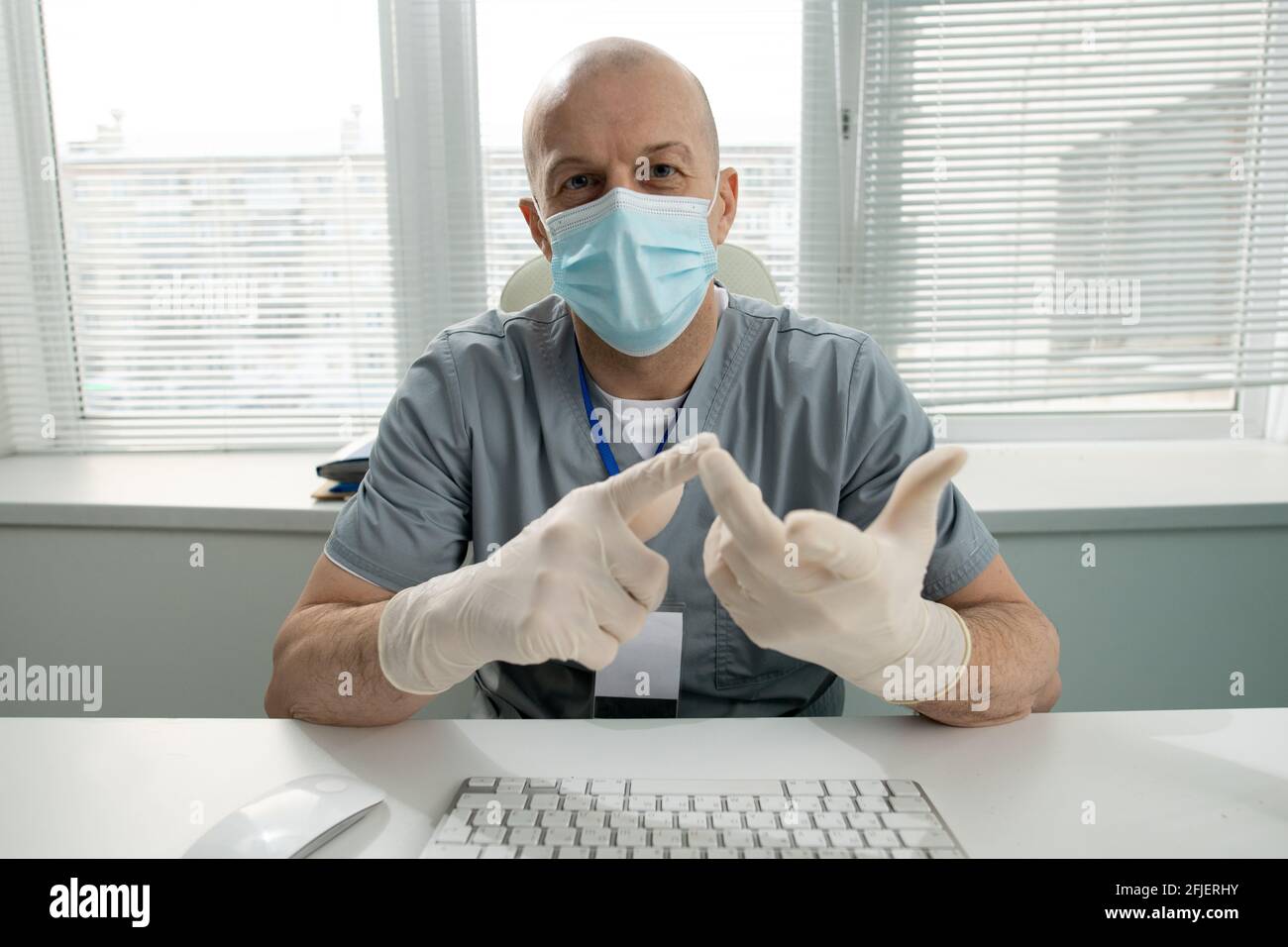 Portrait d'un médecin bald d'âge moyen dans un masque et des gants assis au tableau et compte les doigts tout en énumérant les symptômes Banque D'Images