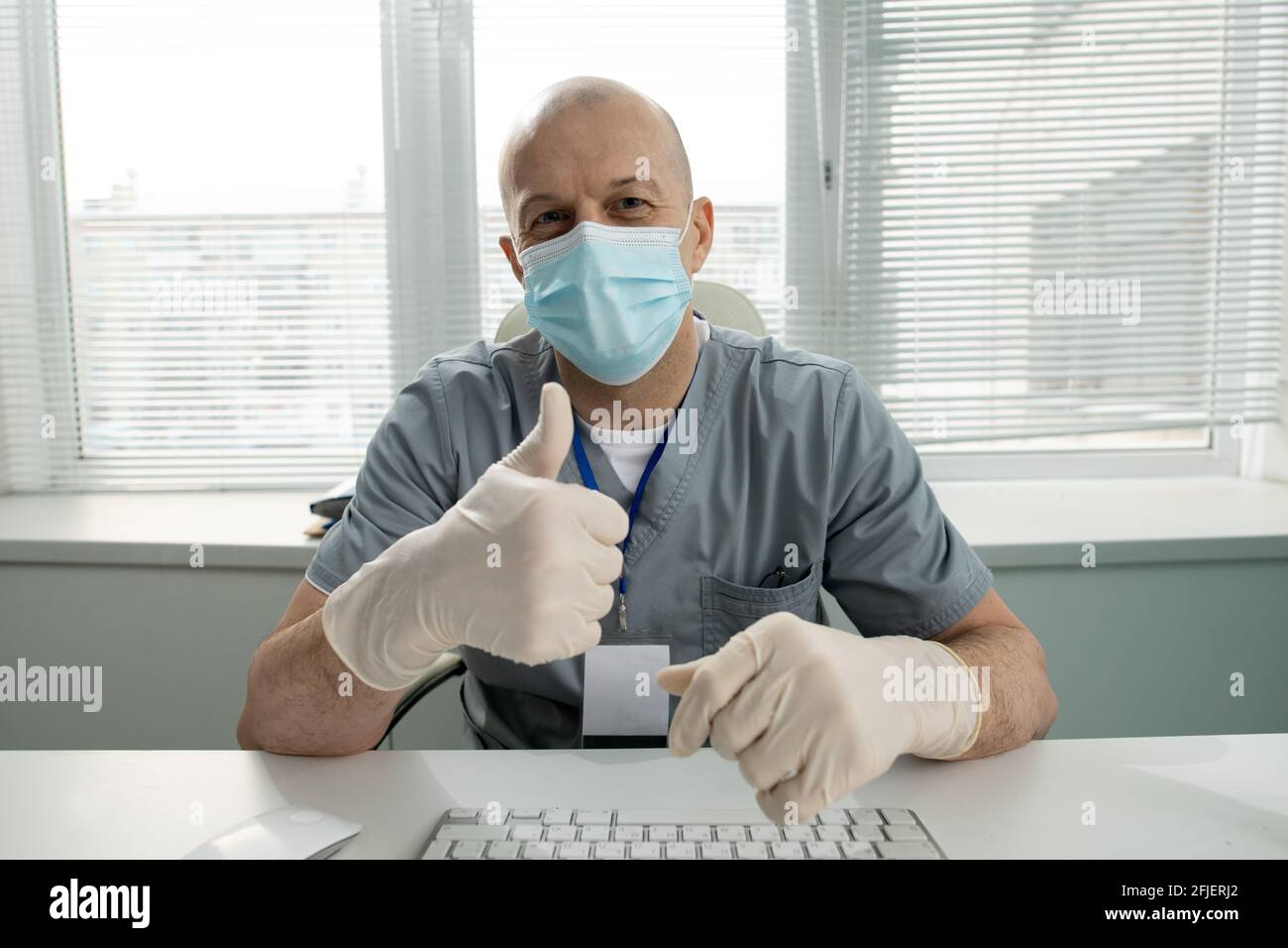 Portrait d'un médecin chauve satisfait en masque facial et en latex gants assis à la table et montrant le pouce vers le haut Banque D'Images