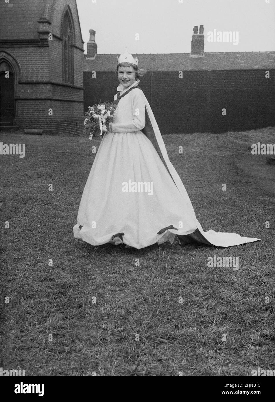 1956, historique, à l'extérieur dans le domaine de l'église, debout pour sa photo, la Reine de mai de la ville, une jeune fille portant une couronne et une longue robe, avec bouquet de fleurs. Angleterre, Royaume-Uni. Un ancien festival célébrant l'arrivée du printemps, le jour de mai a impliqué le couronnement d'une reine de mai et la danse autour d'un Maypole, des activités qui ont eu lieu en Angleterre pendant des siècles. Sélectionné parmi les filles de la région, la Reine de mai commencerait la procession des chars et de la danse. Dans le Nord industrialisé de l'Angleterre, les écoles du Dimanche de l'Église dirigeaient souvent son organisation. Banque D'Images