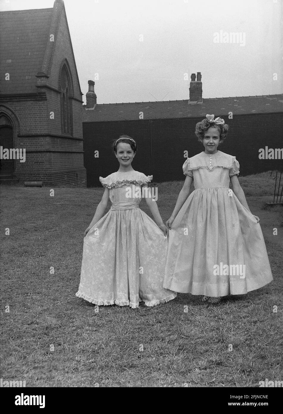 1956, historique, à l'extérieur dans le domaine de l'église, deux jeunes filles montrent leurs jolies robes pour le carnaval du jour de mai, où ils souserviront la Reine de mai de l'année, Leeds, Angleterre, Royaume-Uni. Un ancien festival célébrant l'arrivée du printemps, le jour de mai a impliqué le couronnement d'une reine de mai et la danse autour d'un Maypole, des activités qui ont eu lieu en Angleterre pendant des siècles. Sélectionné parmi les filles de la région, la Reine de mai commencerait la procession des chars et de la danse. Dans le Nord industrialisé de l'Angleterre, les écoles du dimanche de l'Église ont souvent dirigé l'organisation. Banque D'Images
