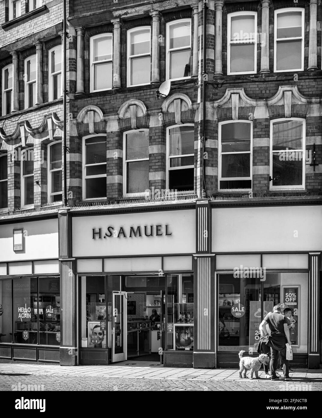 Un magasin sous un bâtiment en briques orné avec des rangées de fenêtres. Une famille avec un chien est d'un côté et les portes du magasin sont ouvertes. Banque D'Images