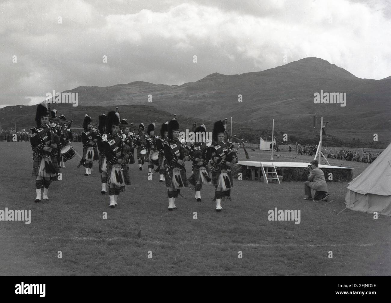1956, historique, un groupe de tubes écossais jouant aux jeux de Greenock Highland, Écosse. Tout d'abord documentés dans les années 1820 à Invergarry, les jeux verront les habitants démontrer leur force et leur agilité dans diverses activités telles que lancer le marteau, ils comprenaient également la danse écossaise et la tuyauterie. La visite de la reine Victoria au rassemblement de braemer en 1848 a commencé l'Association royale et les a vu augmenter leur popularité. Divers événements, connus sous le nom de Highland Games ou simplement des rassemblements, se tiennent maintenant dans toute l'Écosse pendant les mois d'été. Banque D'Images