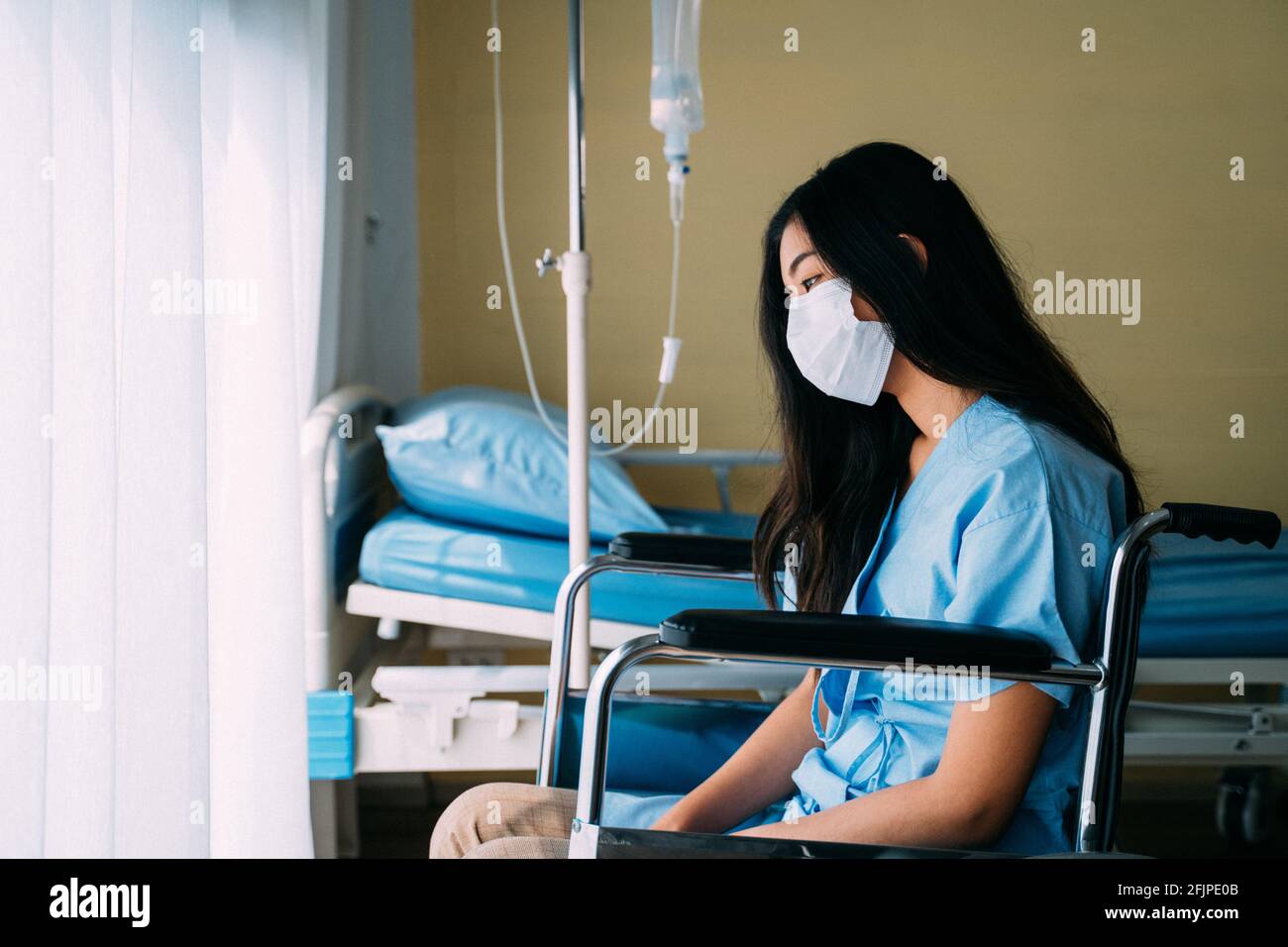 Jeune patiente triste et bouleversée portant un masque chirurgical assis enfoncé sur le fauteuil roulant dans la chambre d'hôpital à côté de la fenêtre Banque D'Images