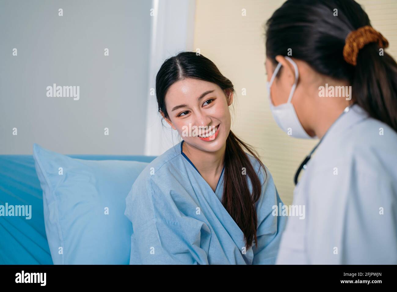 Portrait de la belle jeune femme asiatique en exfoliant parler au médecin portant un masque pour la sécurité lorsqu'il est assis lit d'hôpital pendant la consultation Banque D'Images