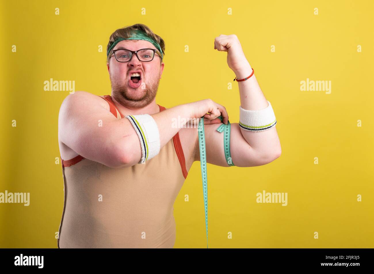 Un drôle, gai gros homme dans les vêtements de sport mesure ses bras. L'homme en surpoids entre pour le sport. Arrière-plan jaune isolé Banque D'Images