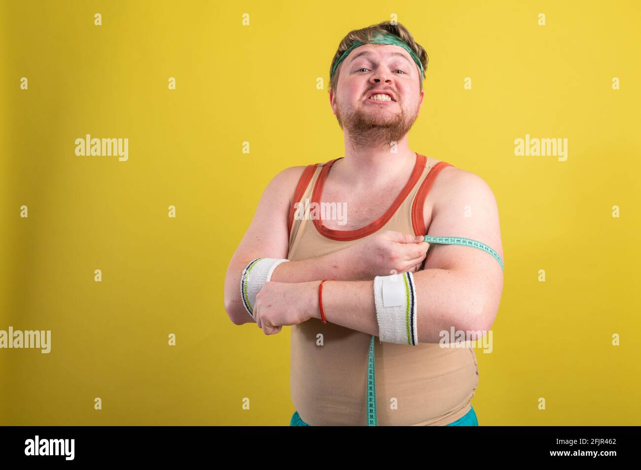 Un drôle, gai gros homme dans les vêtements de sport mesure ses bras. L'homme en surpoids entre pour le sport. Arrière-plan jaune isolé Banque D'Images