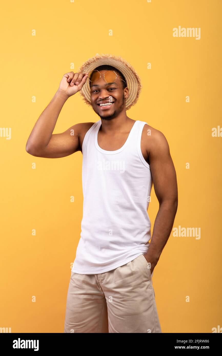 Des vibes d'été. Portrait d'un jeune homme noir en t-shirt, short, chapeau de paille et lunettes de soleil posant et souriant à l'appareil photo Banque D'Images