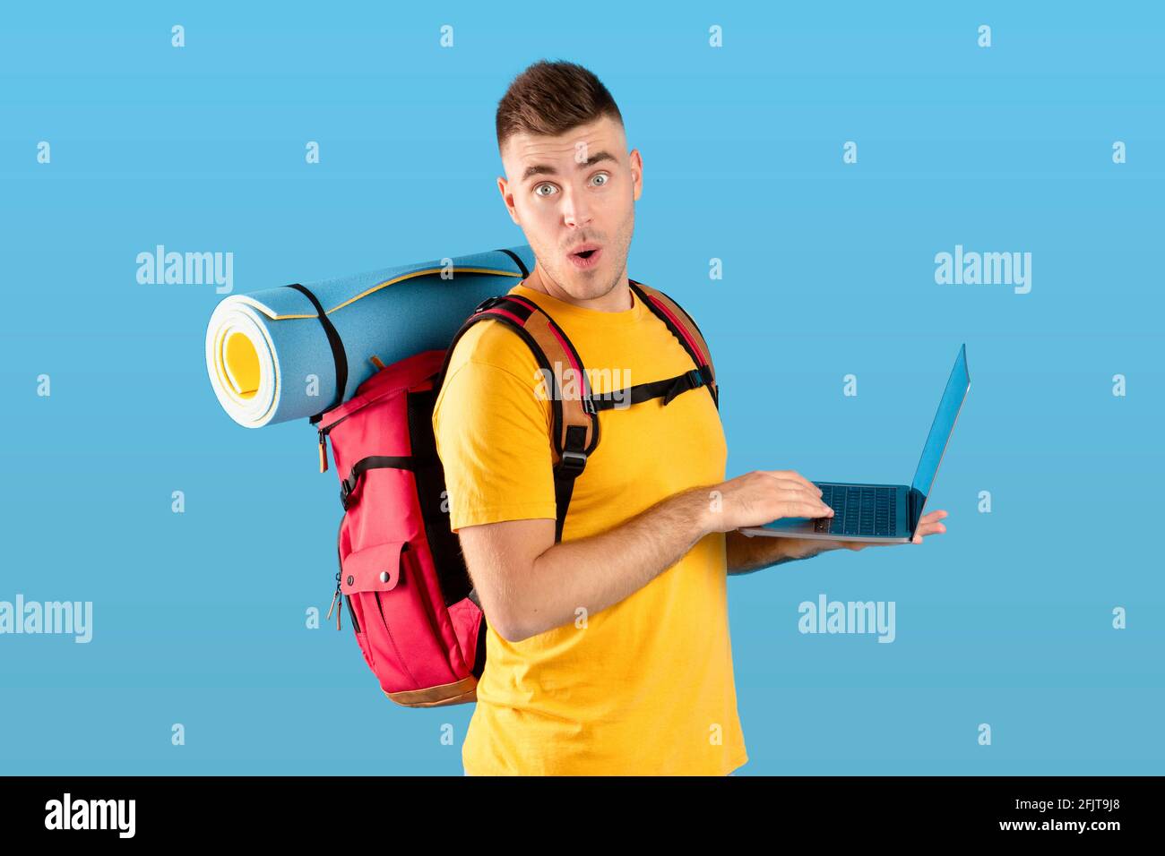 Réservation de la visite en ligne. Jeune touriste avec ordinateur portable surpris par une grande réduction dans l'agence de voyage en ligne, fond bleu Banque D'Images