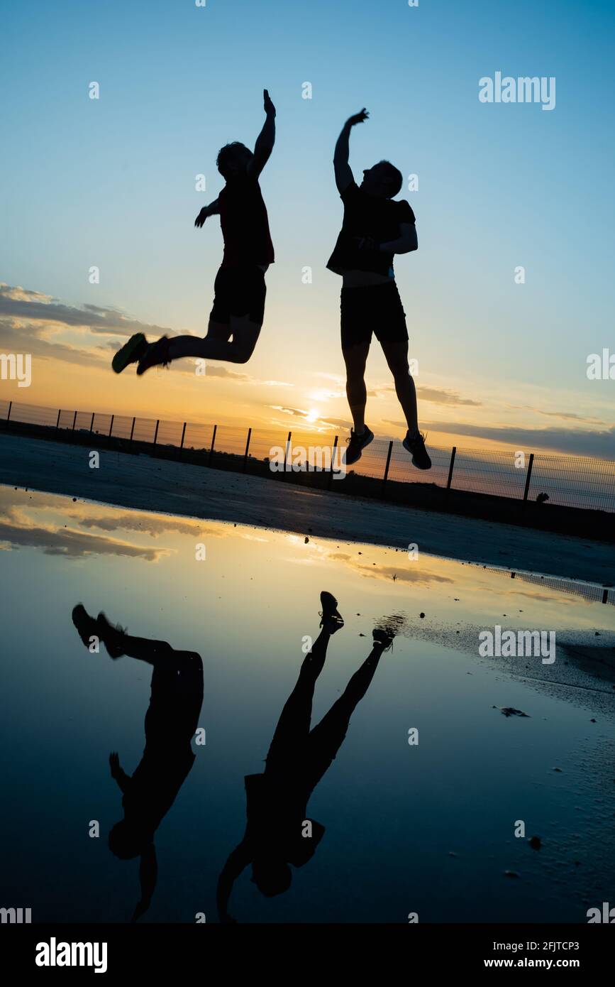 Concept: Sport. Deux sportifs en plein air. Silhouette de deux hommes méconnaissables qui sautent en hauteur. Coucher de soleil avec ciel épique. Réflexion dans l'eau Banque D'Images