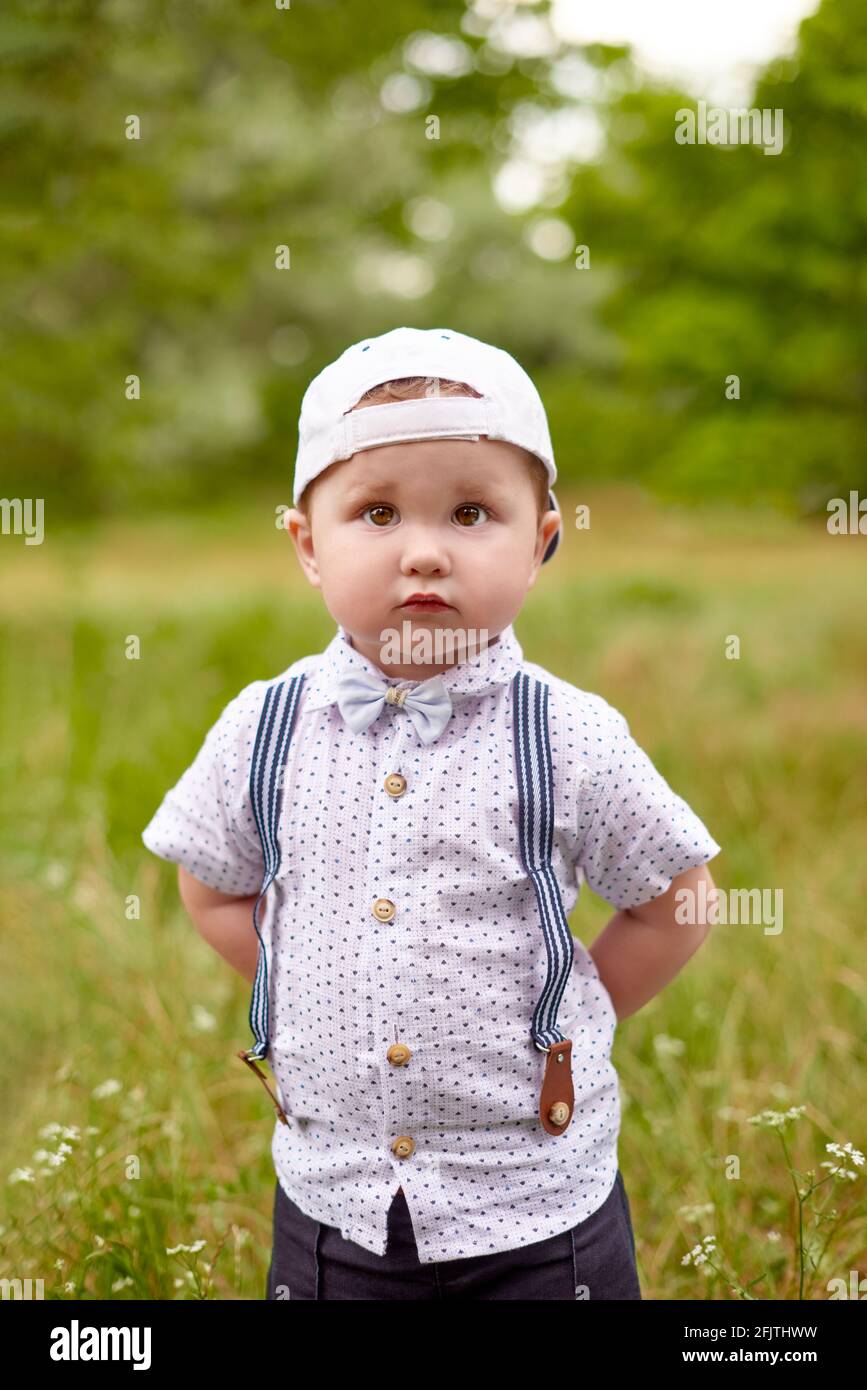 petit garçon sérieux dans des bretelles et une casquette lui cache les  mains derrière le dos en position de stationnement Photo Stock - Alamy
