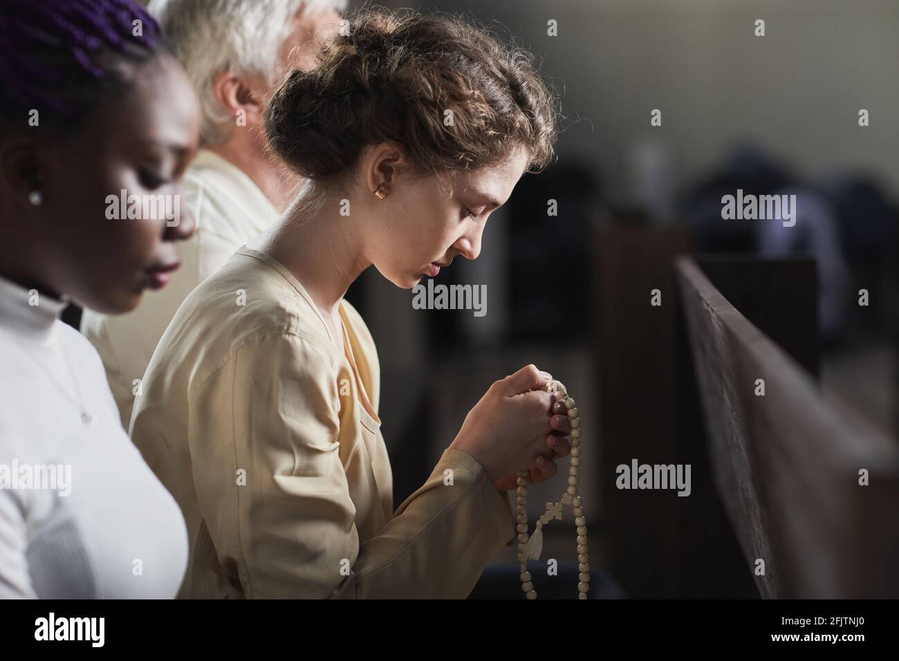 Jeune femme tenant rosaire tout en priant dans l'église parmi autres personnes Banque D'Images