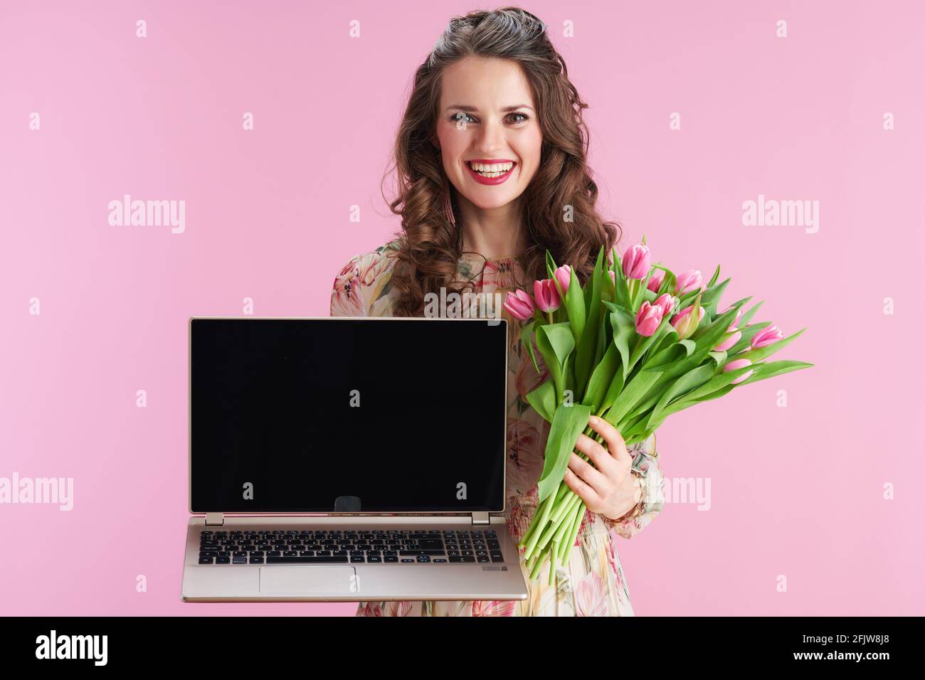 femme moderne souriante en robe florale avec bouquet de tulipes montrant ordinateur portable écran vierge sur fond rose. Banque D'Images