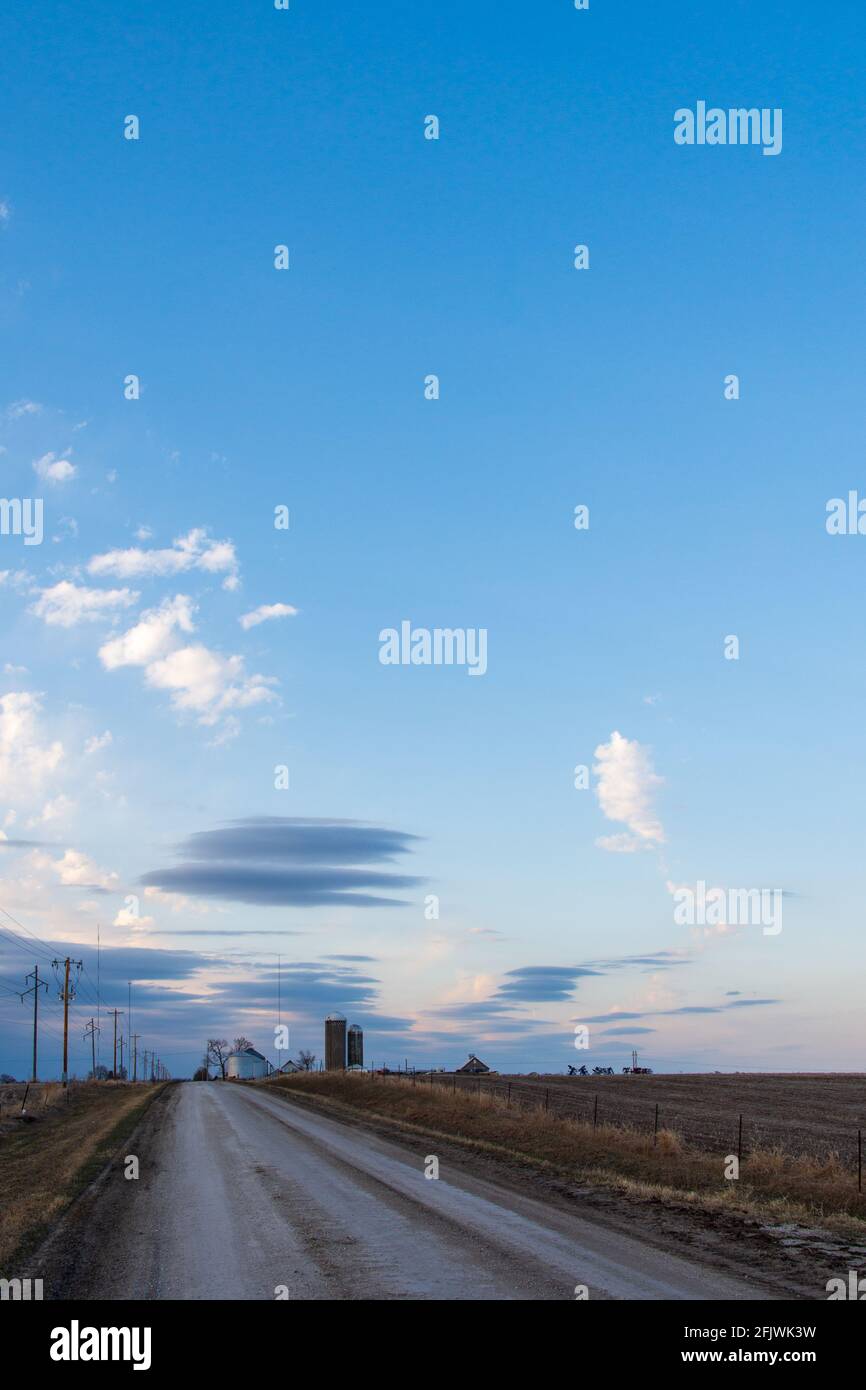 Ciel bleu et nuages du soir au-dessus d'une route de campagne en gravier avec des silos de ferme et des silos à grains éloignés. Banque D'Images