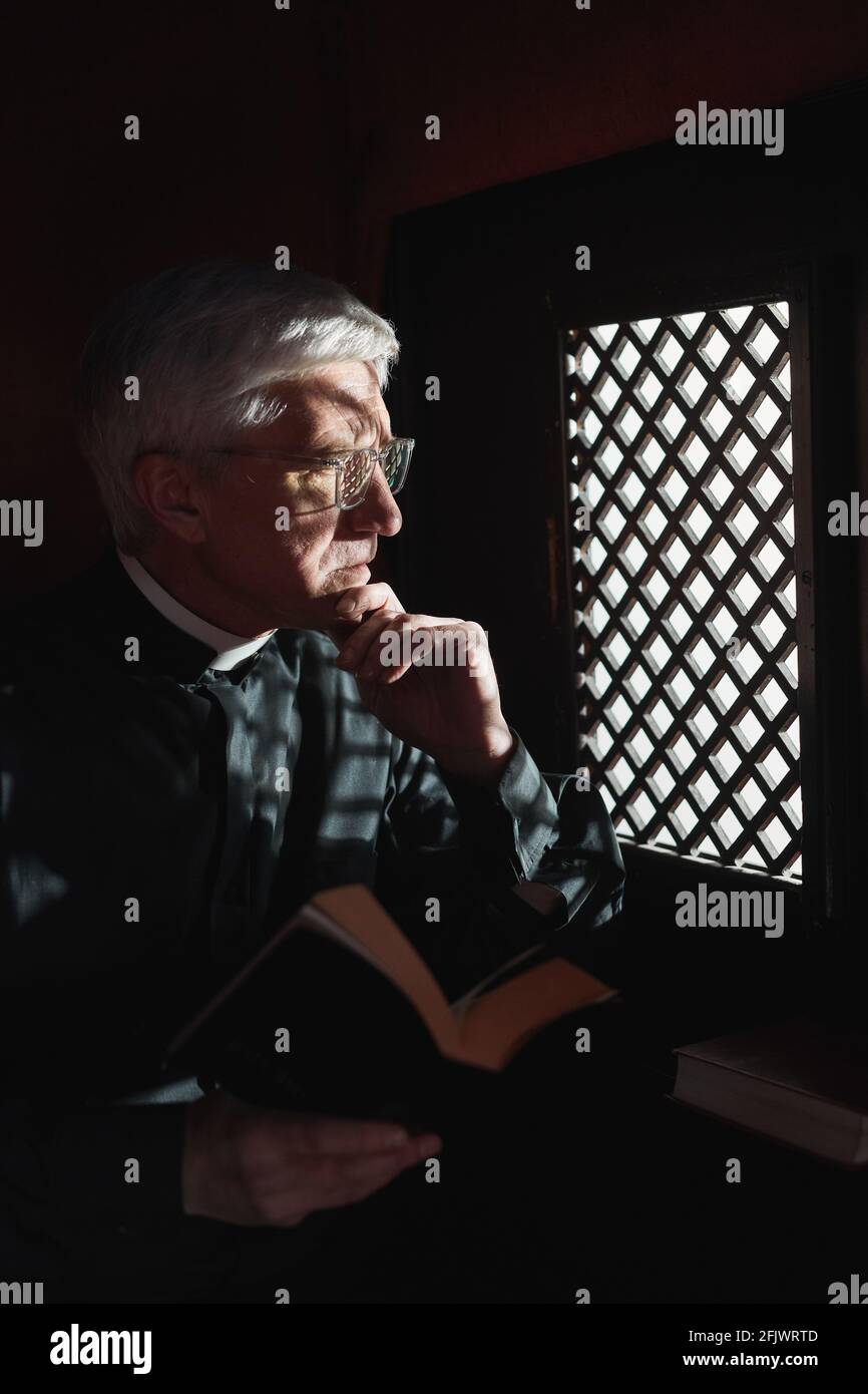 Prêtre senior sérieux assis avec la Bible en confessionnel dans le église Banque D'Images