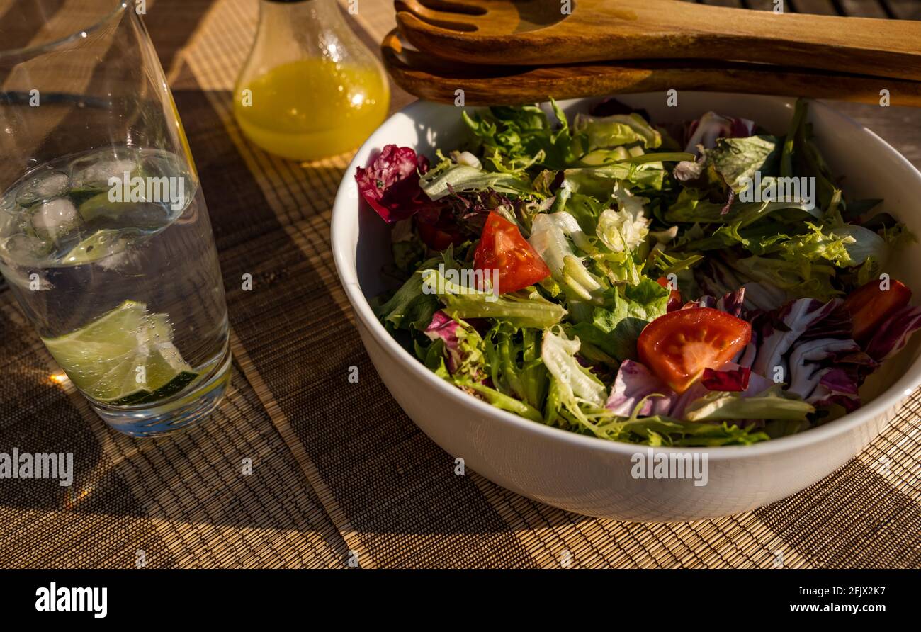 Bol à salade avec légumes verts, laitue et tomates, salade en bois, cuillères à servir sur une table extérieure au soleil, avec une bouteille de vinaigrette Banque D'Images