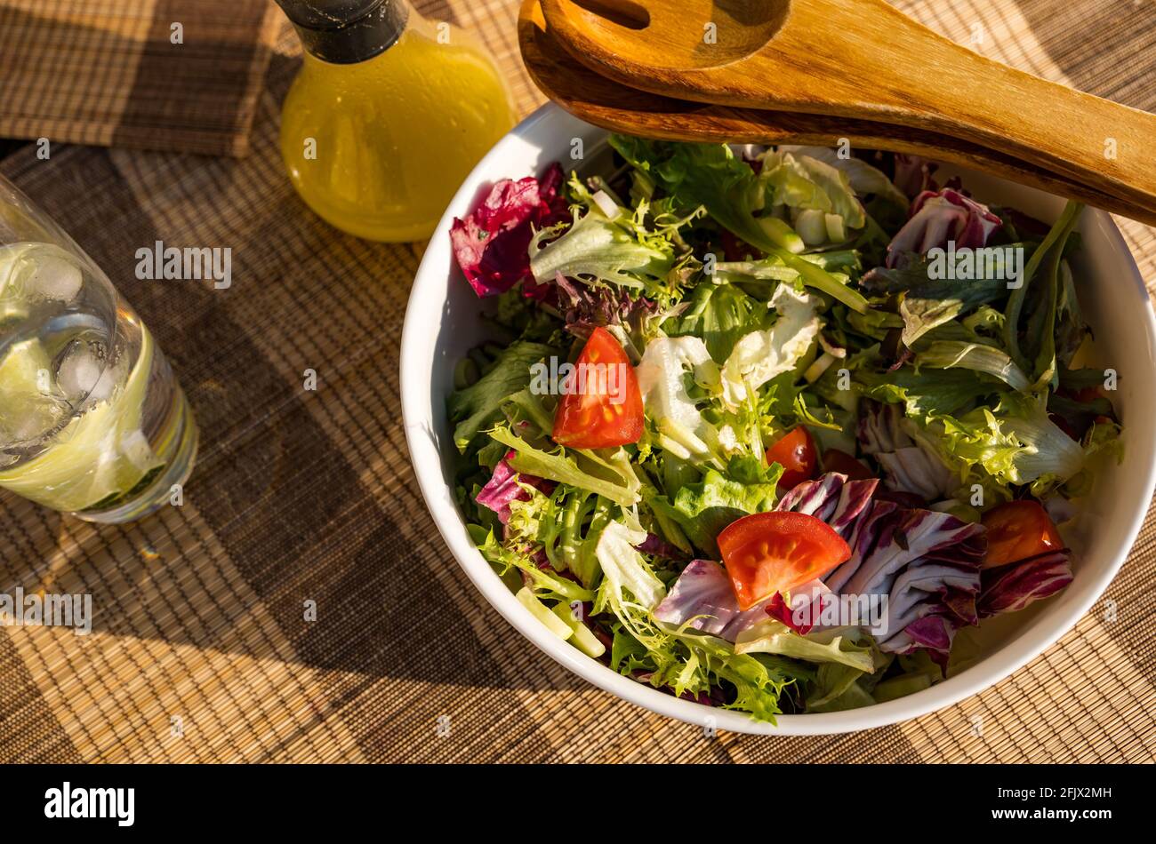 Bol à salade avec légumes verts, laitue et tomates, salade en bois, cuillères à servir sur une table extérieure au soleil, avec une bouteille de vinaigrette Banque D'Images