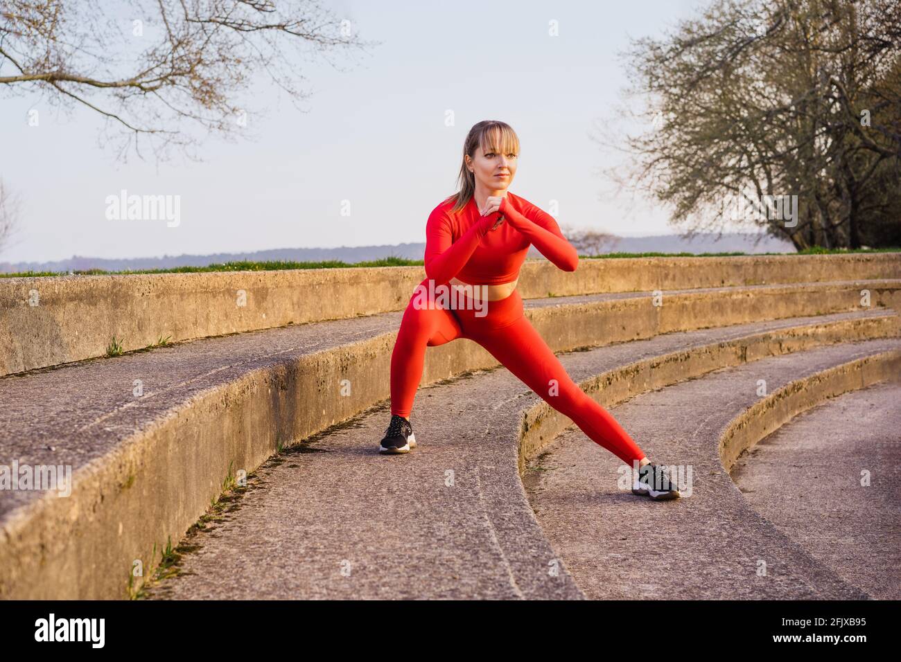 Jeune femme de fitness en rouge sport porter faire une fente latérale, squats pendant l'entraînement en plein air sur les marches du stade. Étirement après l'entraînement. Actif Banque D'Images