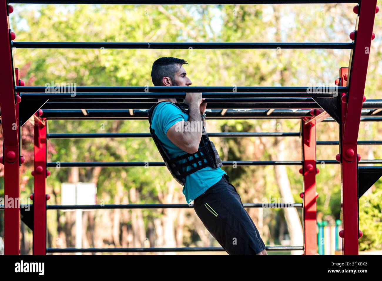 Homme aux cheveux sombres avec barbe faisant un pull-up sur la barre de calisthénique avec gilet de poids. Concept de fitness en plein air. Banque D'Images