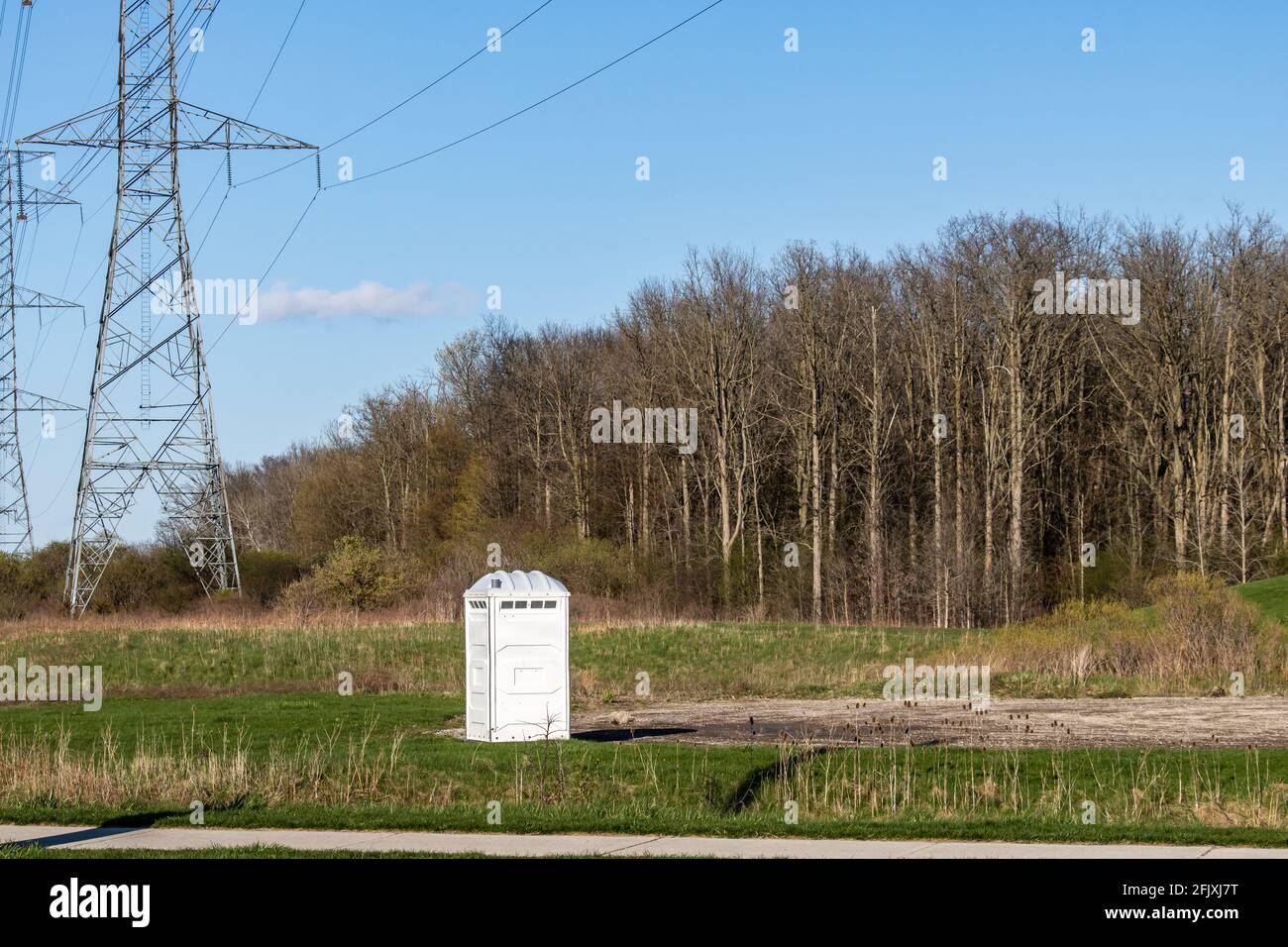 toilette portable blanche au bout d'un parking et à côté d'un champ et de tours hydro-électriques. Forêt en arrière-plan. Banque D'Images