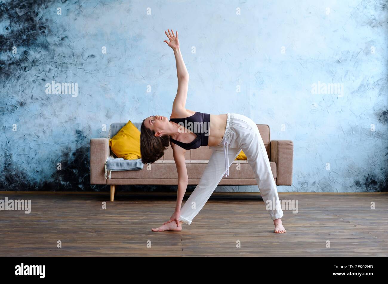Calme jeune femme brunette en blanc utthita trikonasana pratiquant le yoga près du canapé. Banque D'Images