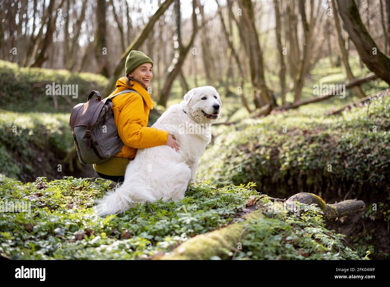 Jeune femme en vêtements de randonnée et sac à dos assis près de grand chien blanc dans la forêt verte de printemps. Aime et explore la nature tranquille. Banque D'Images
