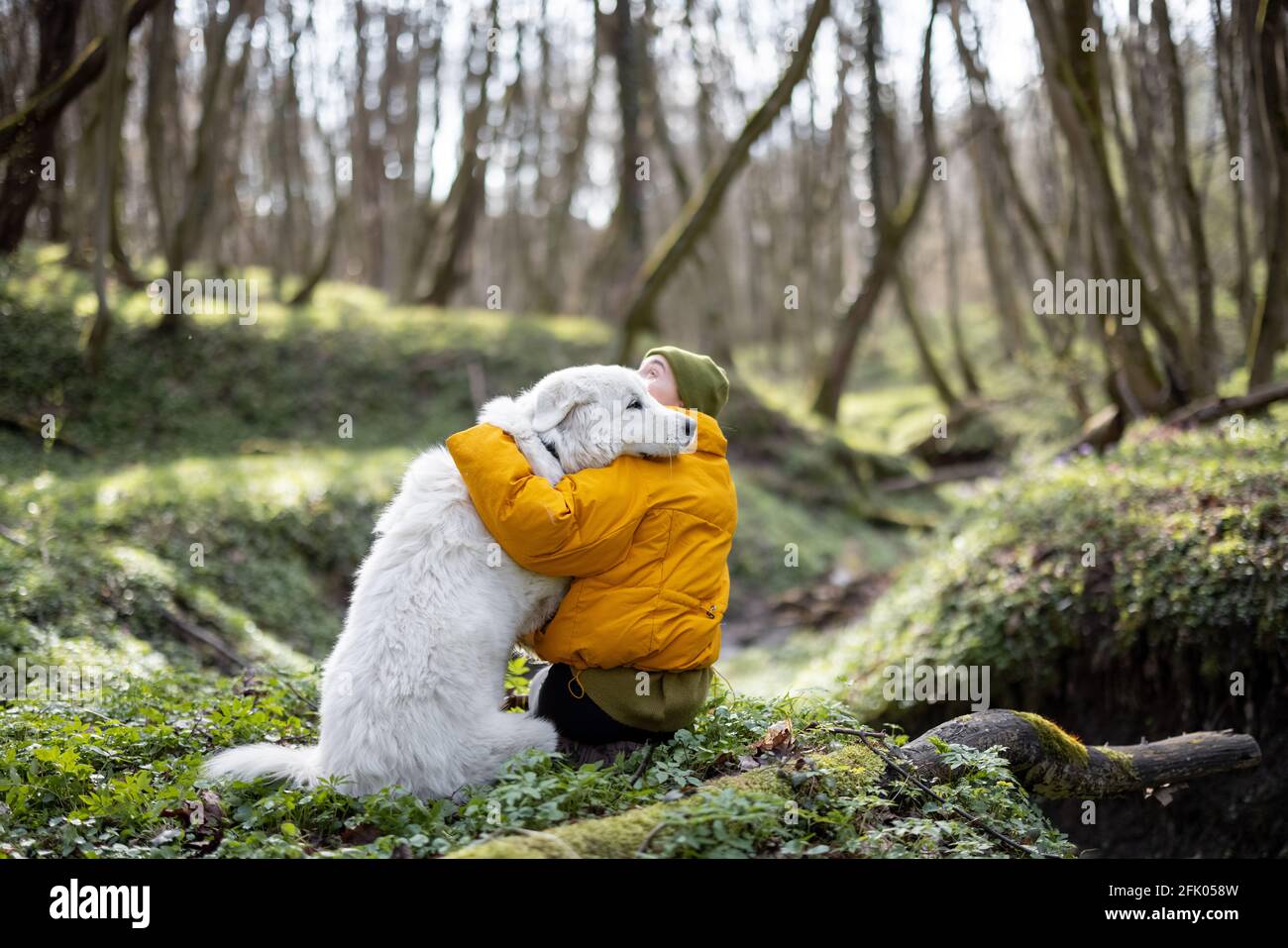 Une jeune femme en vêtements de randonnée, assise près d'un grand chien blanc, encroche son adorable animal de compagnie dans la forêt verte de printemps. Aime et explore la nature tranquille. Banque D'Images