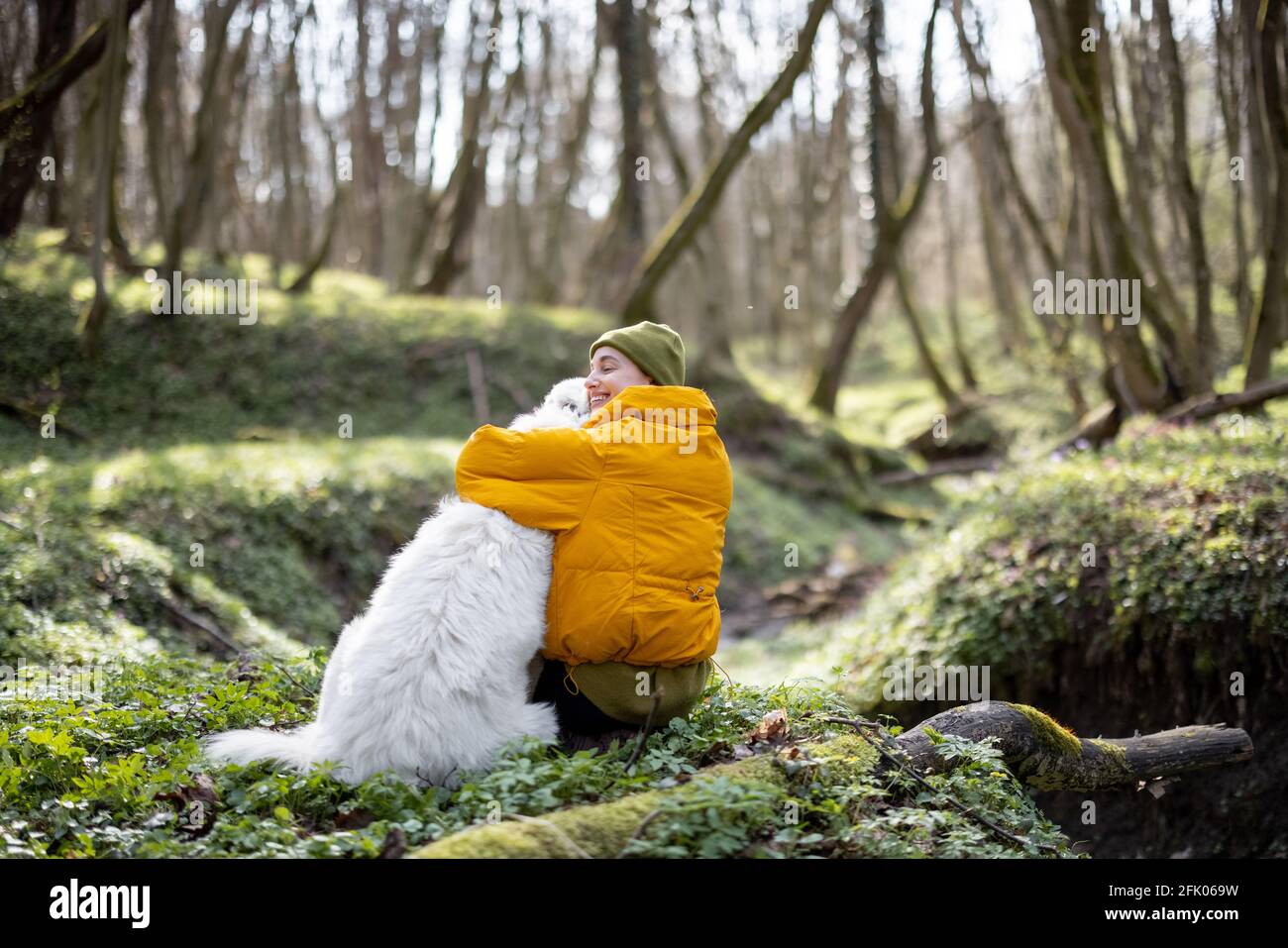 Jeune femme en vêtements de randonnée assis près d'un grand chien blanc et de câlins dans la forêt verte de printemps. Aime et explore la nature tranquille. Banque D'Images