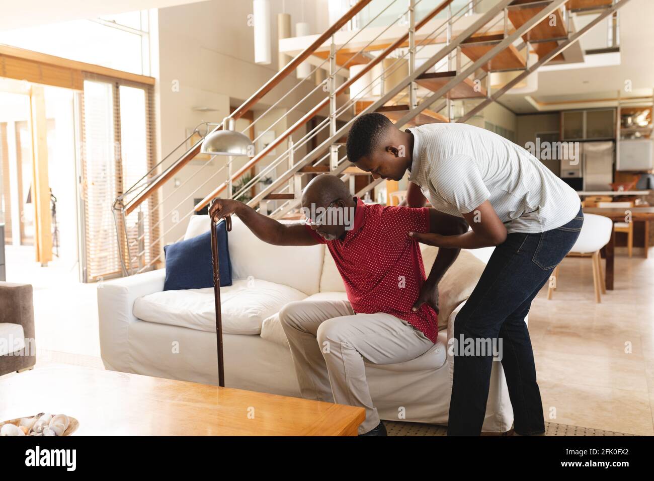 Un jeune homme afro-américain aide son père avec des maux de dos pour sortir du canapé à la maison Banque D'Images