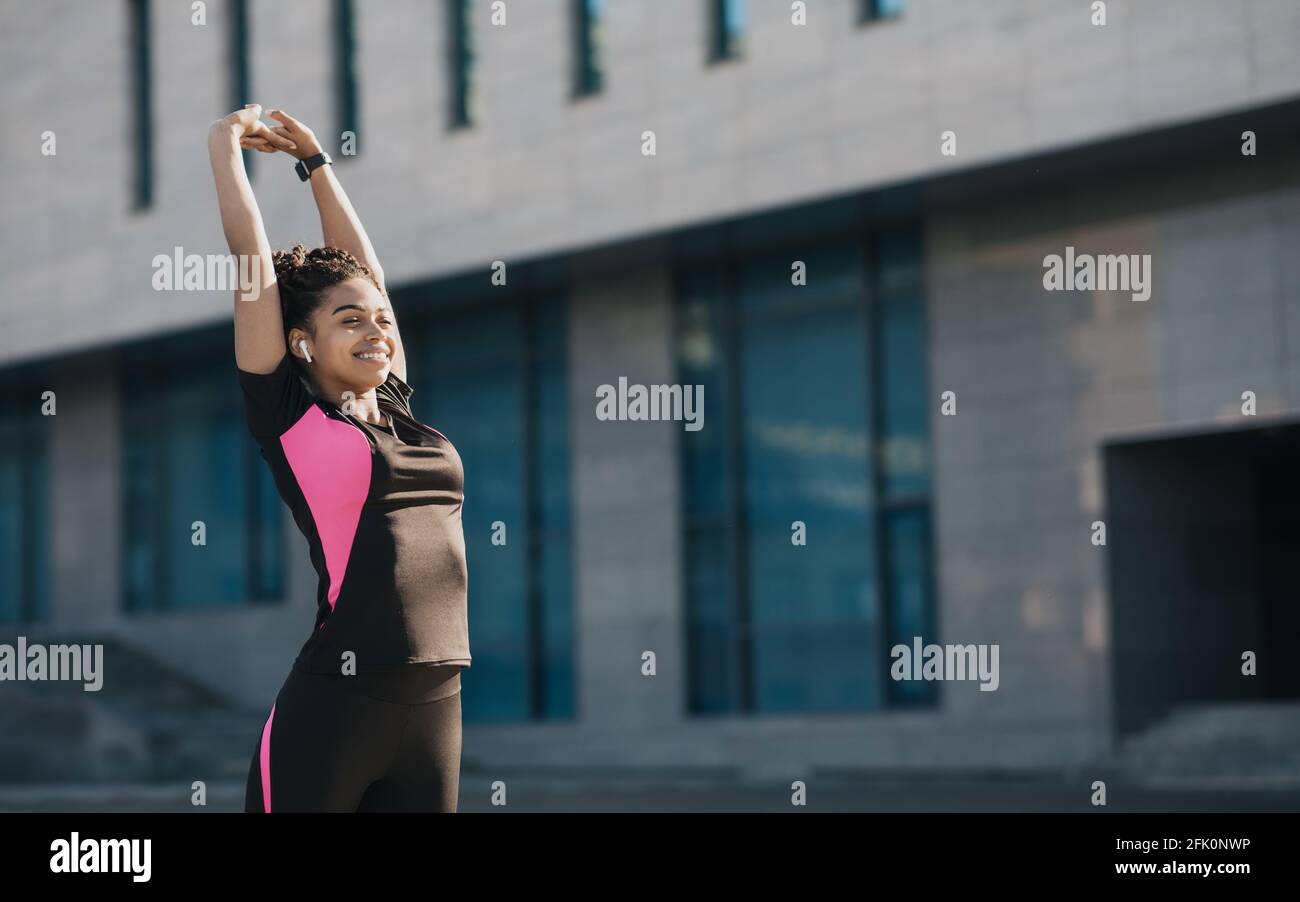 Exercices étirant les muscles du corps en ville, mode de vie sain. Souriant belle jeune femme afro-américaine dans les vêtements de sport et l'entraînement de casque sur m Banque D'Images