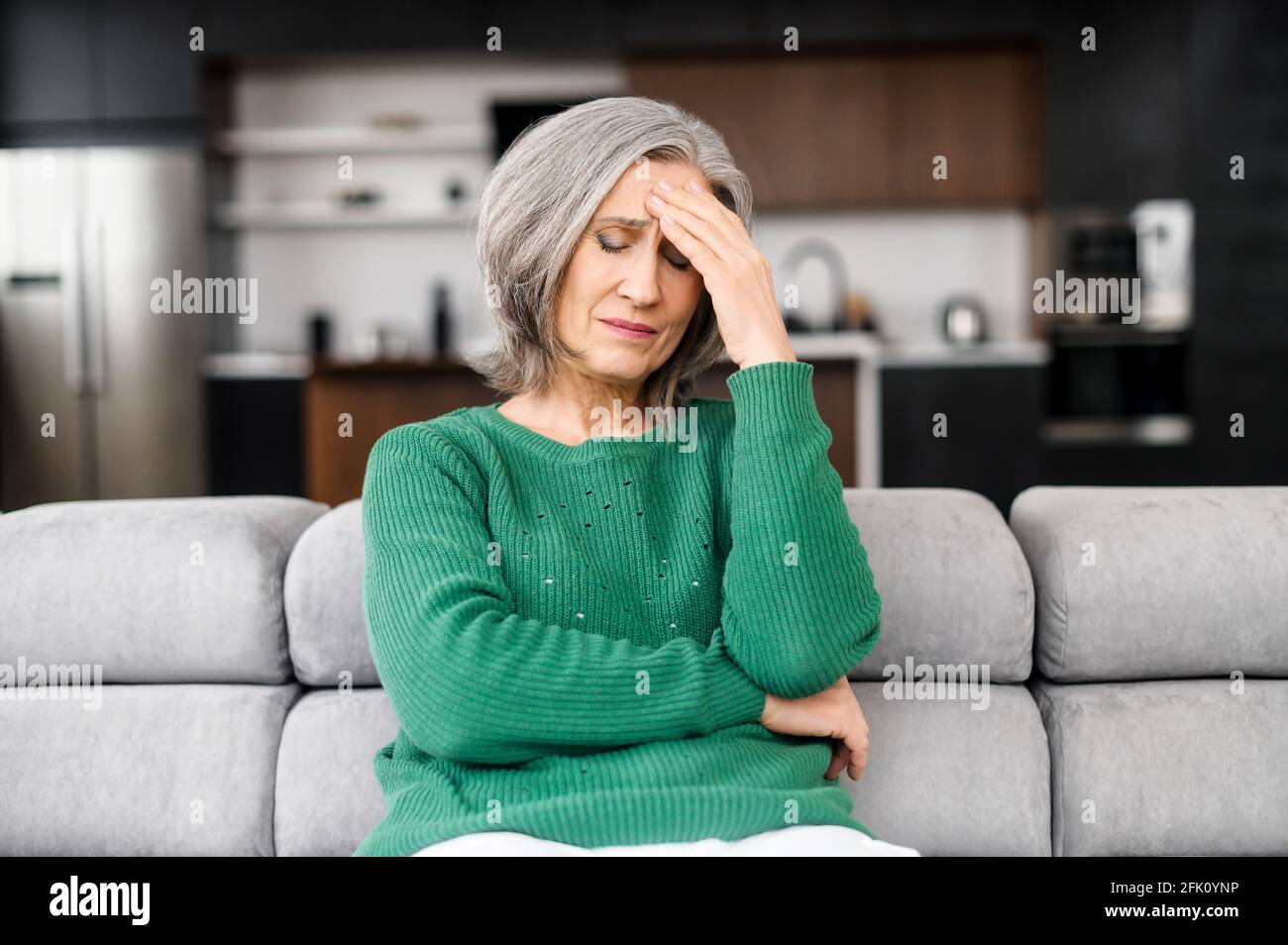 Femme âgée avec des cheveux gris sentir mal de tête, s'assoit sur le canapé à la maison, tient le front avec une expression painée sur son visage, femelle mature souffrant de migraine, se sentant mal Banque D'Images