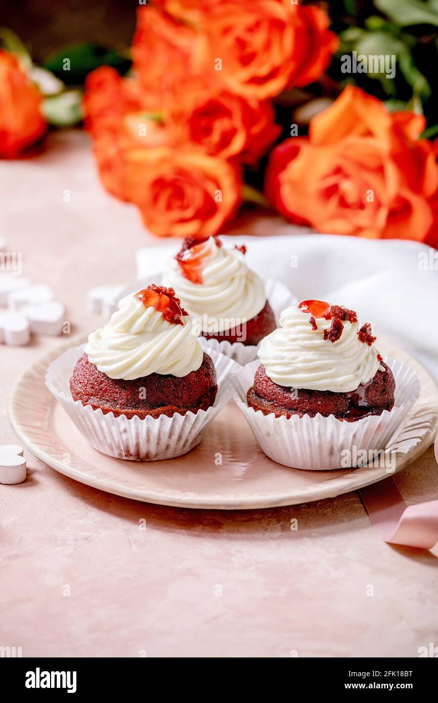 Petits gâteaux faits maison en velours rouge avec crème fouettée sur plaque en céramique rose, serviette blanche avec ruban, fleurs roses, coeurs en bois sur fond de texture rose Banque D'Images