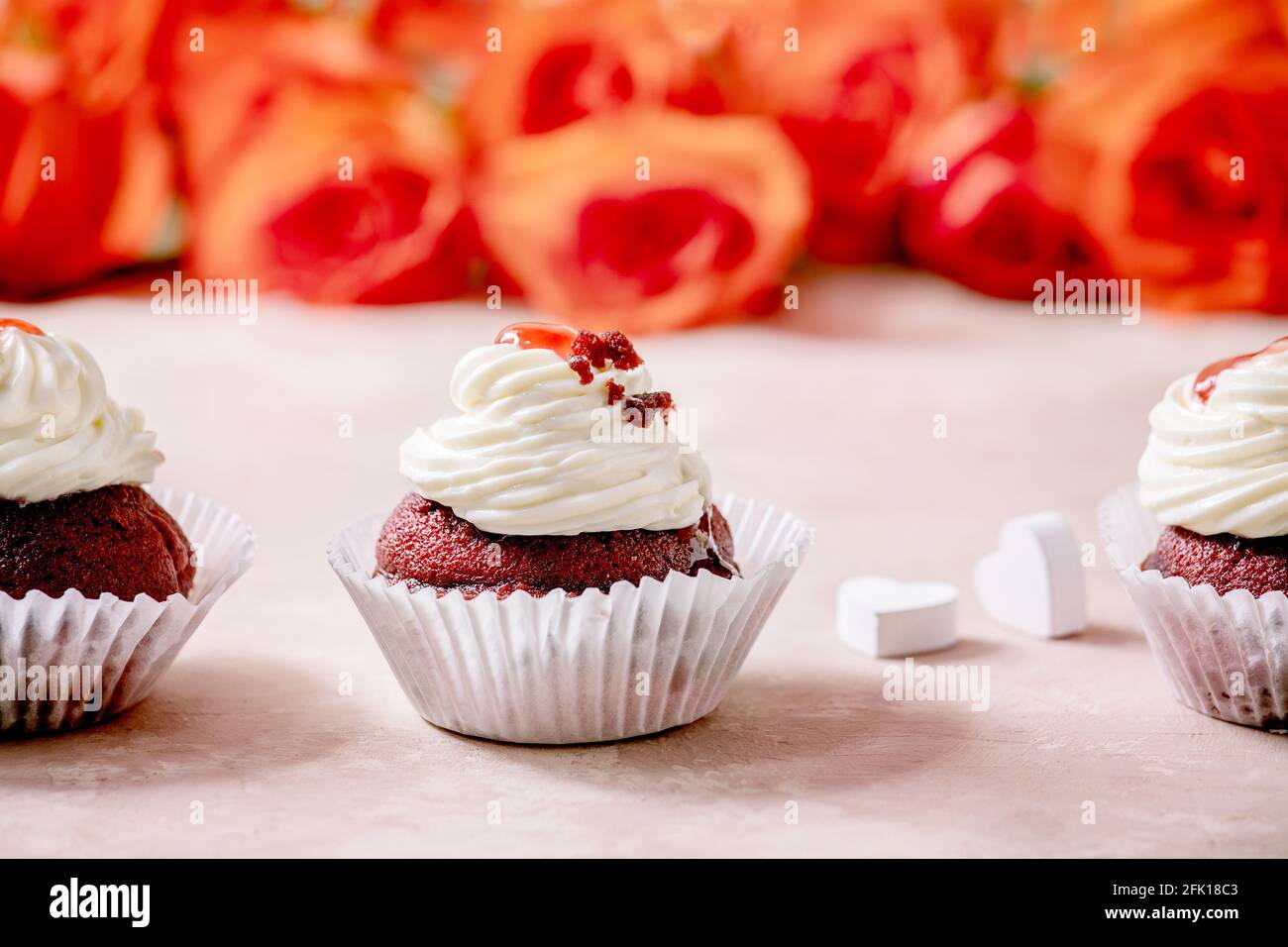 Petits gâteaux faits maison en velours rouge avec crème fouettée en rangée, serviette blanche avec ruban, fleurs roses, coeurs en bois sur fond de texture rose. Saint-Valentin Banque D'Images