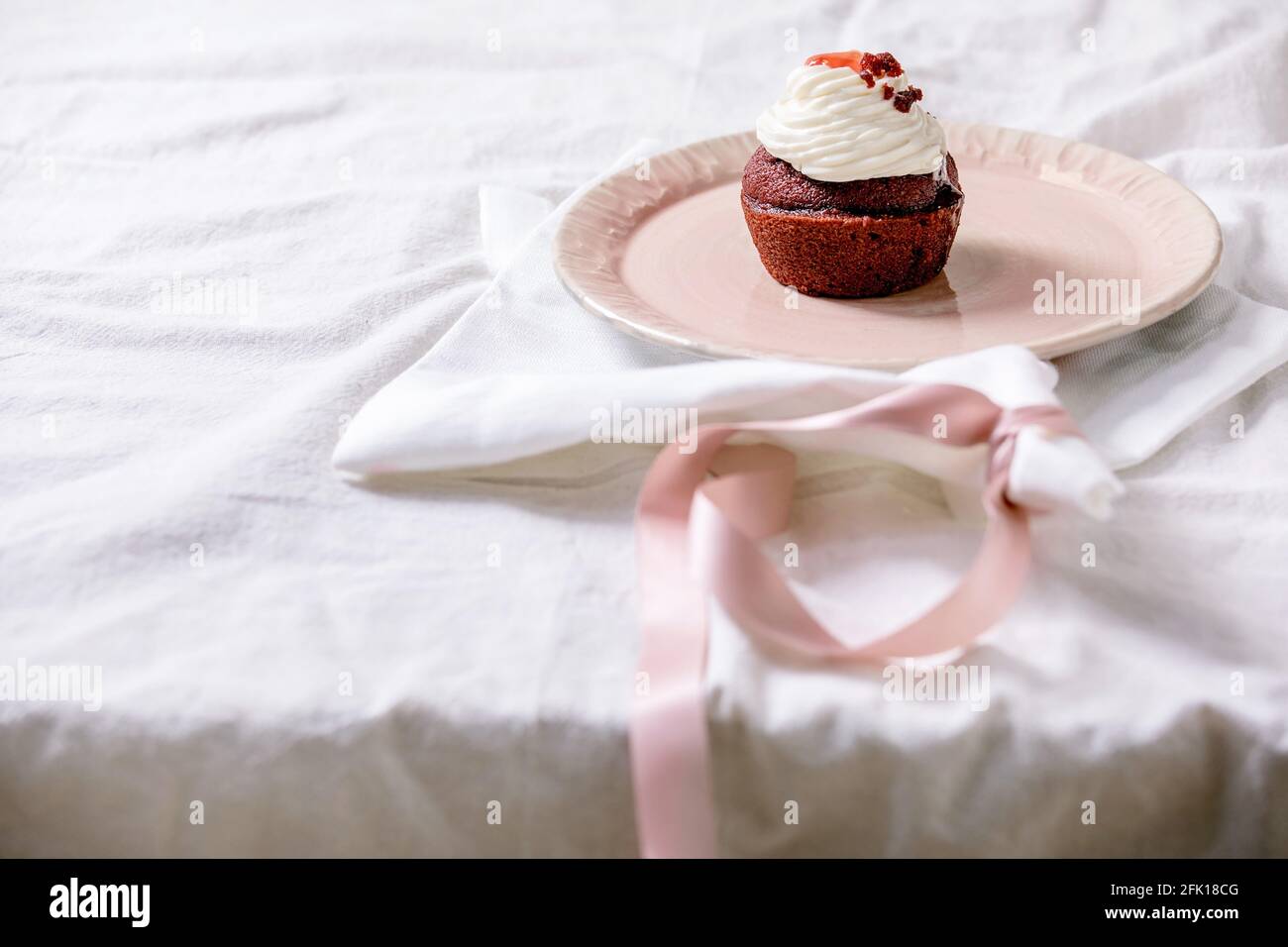 Petit gâteau en velours rouge maison avec crème fouettée sur plaque en céramique rose, serviette blanche avec ruban sur nappe blanche. Copier l'espace Banque D'Images