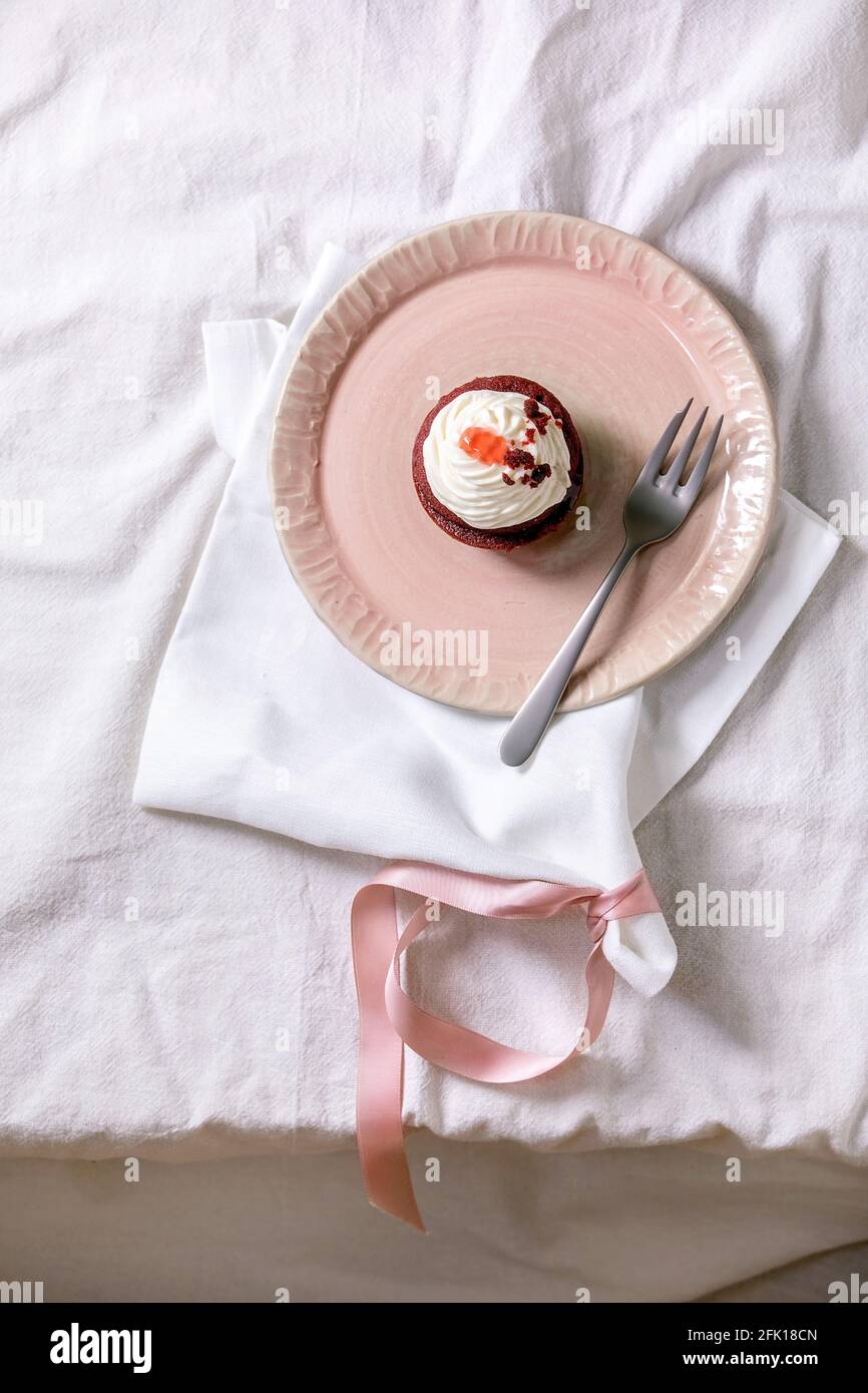 Petit gâteau en velours rouge maison avec crème fouettée sur plaque en céramique rose, serviette blanche avec ruban sur nappe blanche. Pose à plat. Copier l'espace Banque D'Images