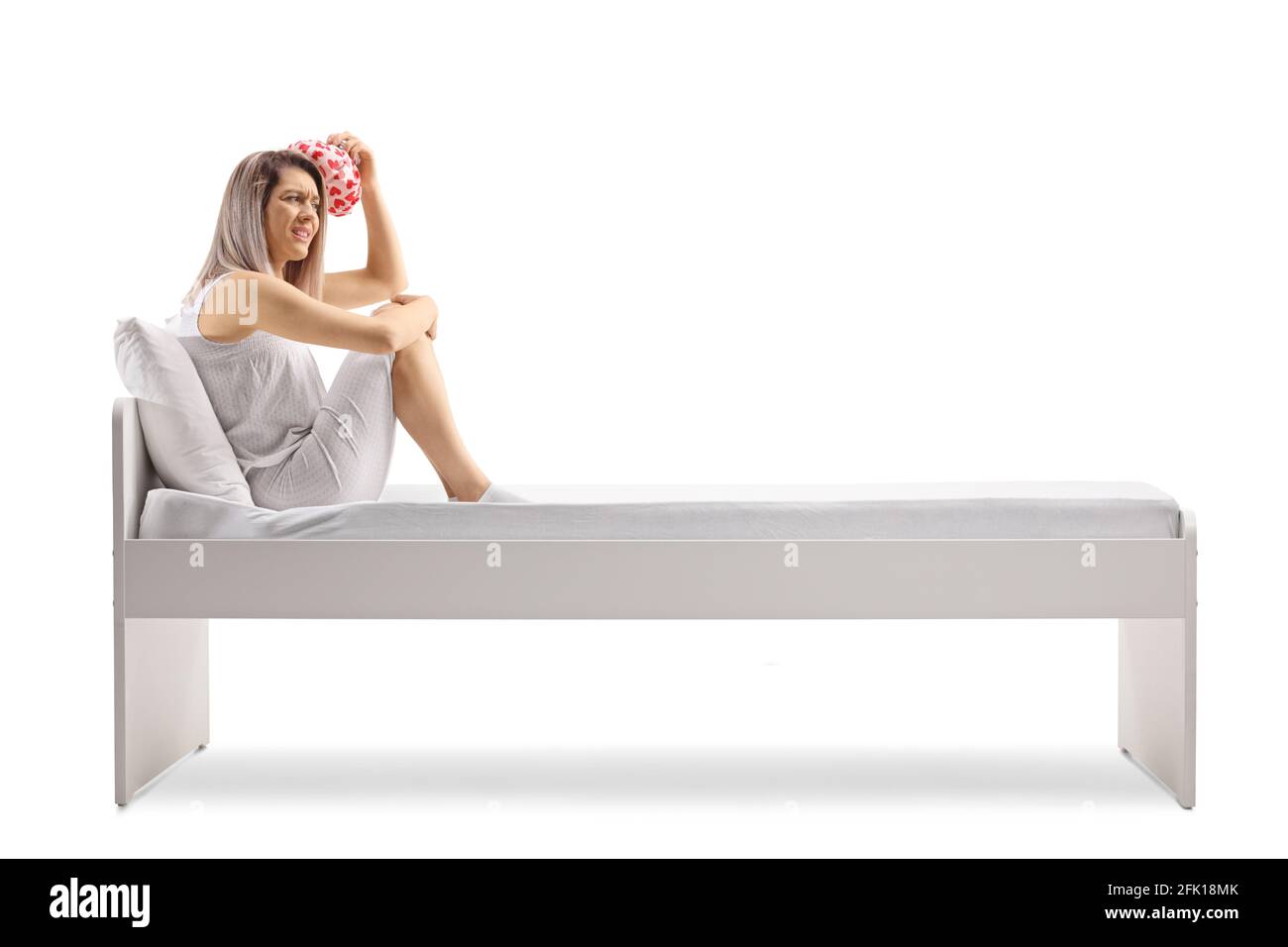 Jeune femme assise sur un lit et tenant un oreiller sur sa tête isolée sur fond blanc Banque D'Images