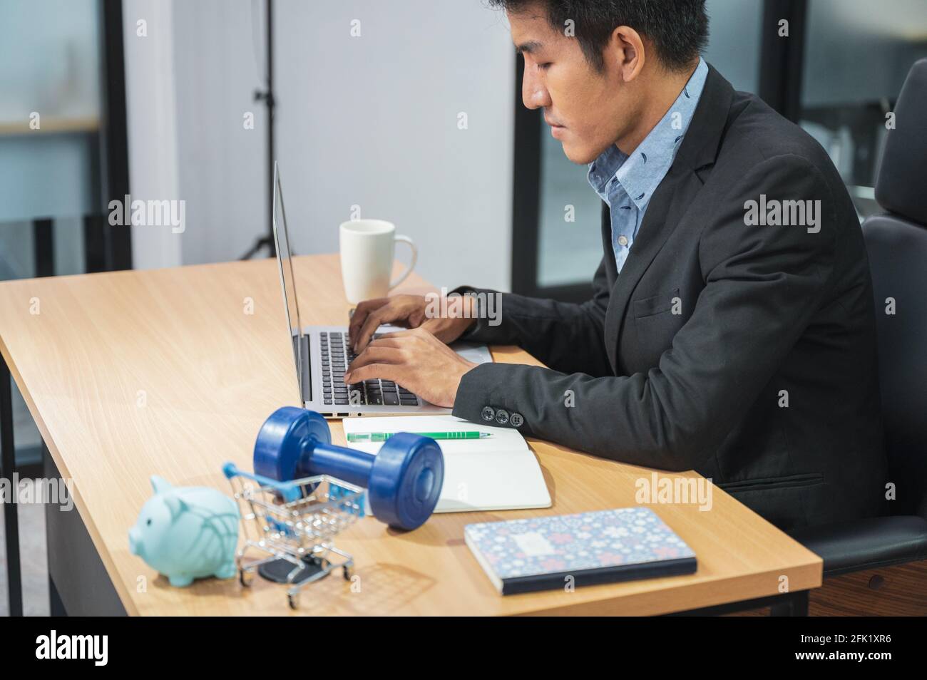 Responsable marketing asiatique homme travaillant avec ordinateur portable et produit piggy banque, dumbell, mini-panier sur le bureau en bois Banque D'Images