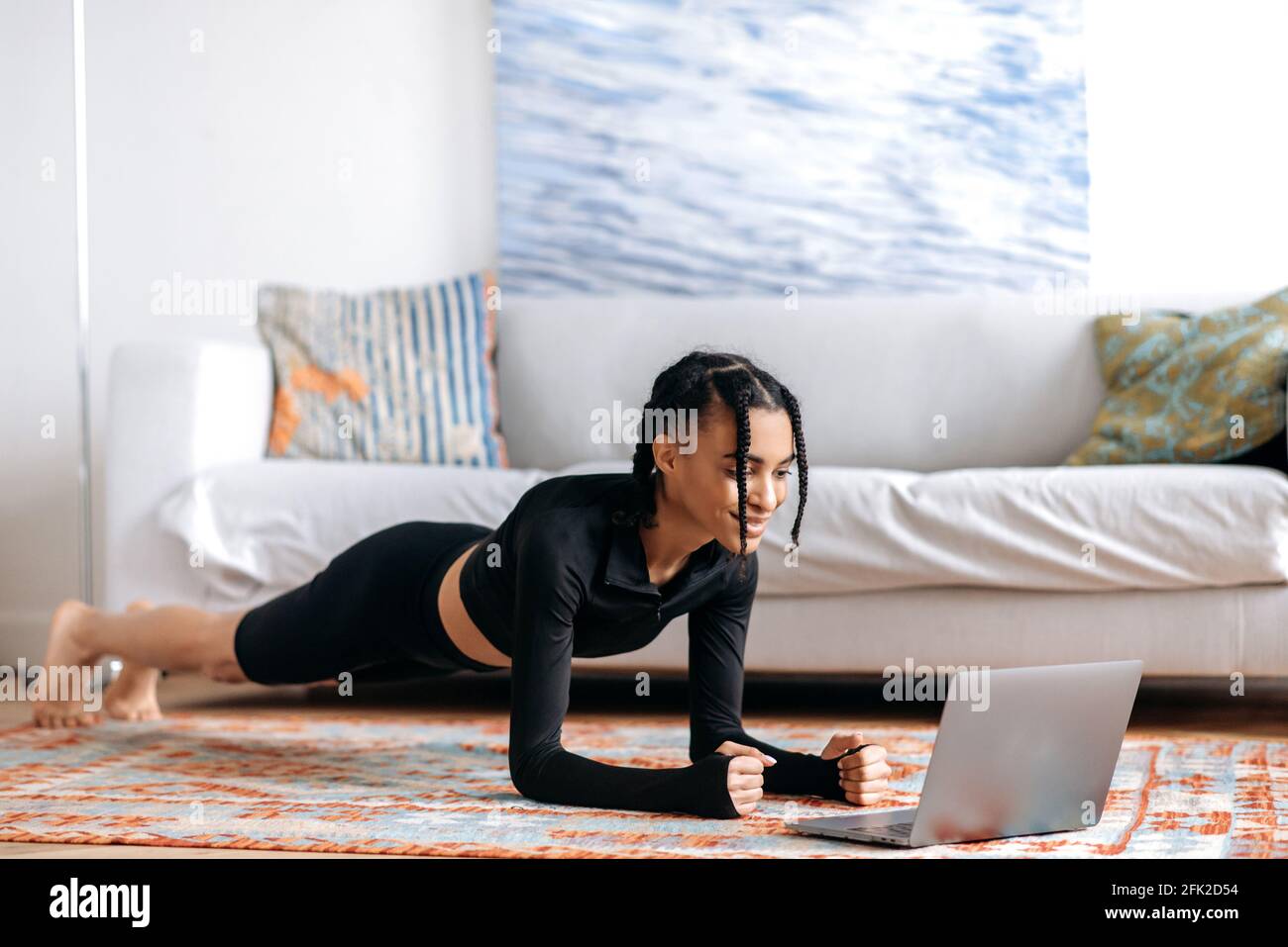 Belle jeune afro-américaine dans les vêtements de sport, faisant une planche sur le sol à la maison, à l'aide d'un ordinateur portable, regardant une vidéo de fitness ou des cours de yoga, menant un style de vie sain, souriant Banque D'Images