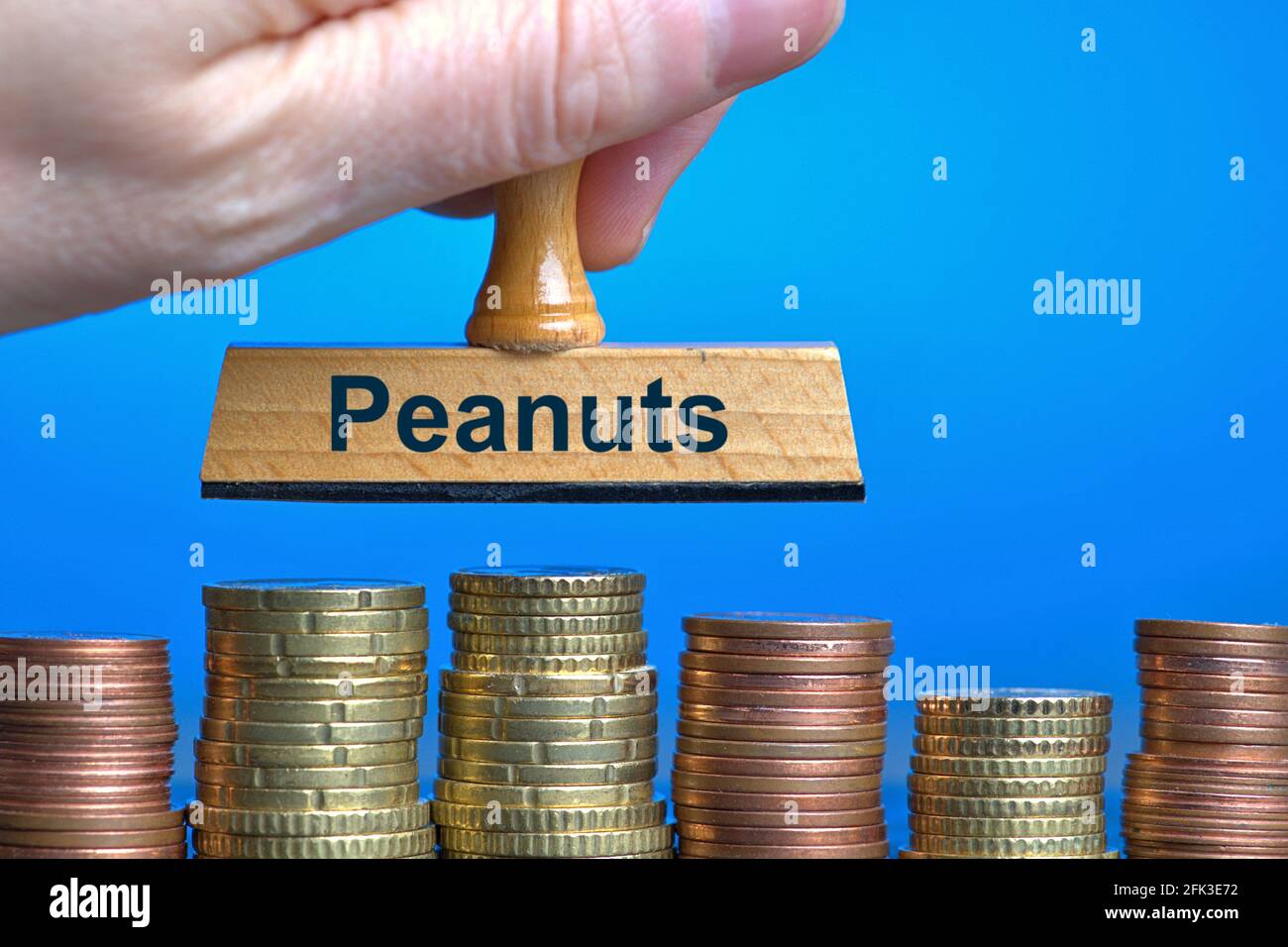 Allemagne. 27 avril 2021. Un timbre en bois avec l'inscription 'Peanuts', tenu à la main dans la coupe de plusieurs piles de pièces en euros/centimes. Photo avec composition sur fond bleu. Les arachides sont familièrement une expression pour de petites choses ou des sommes insignifiantes d'argent. Le mot a été déclaré le mot allemand de l'année 1994 | usage dans le monde crédit: dpa/Alay Live News Banque D'Images