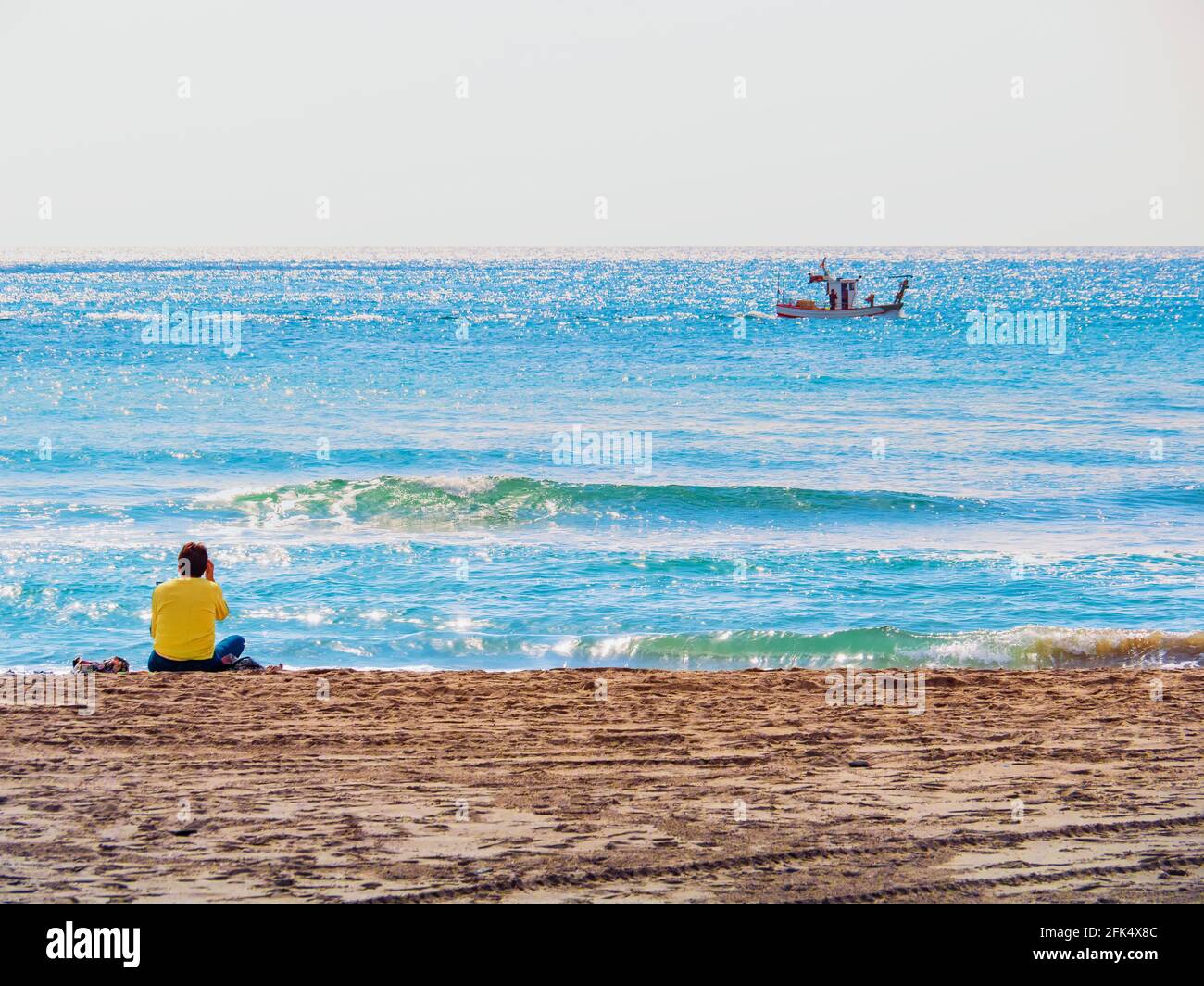 Femme en cavalier jaune assise sur le sable de la plage de Marbella regardant un bateau passant à l'horizon. Banque D'Images