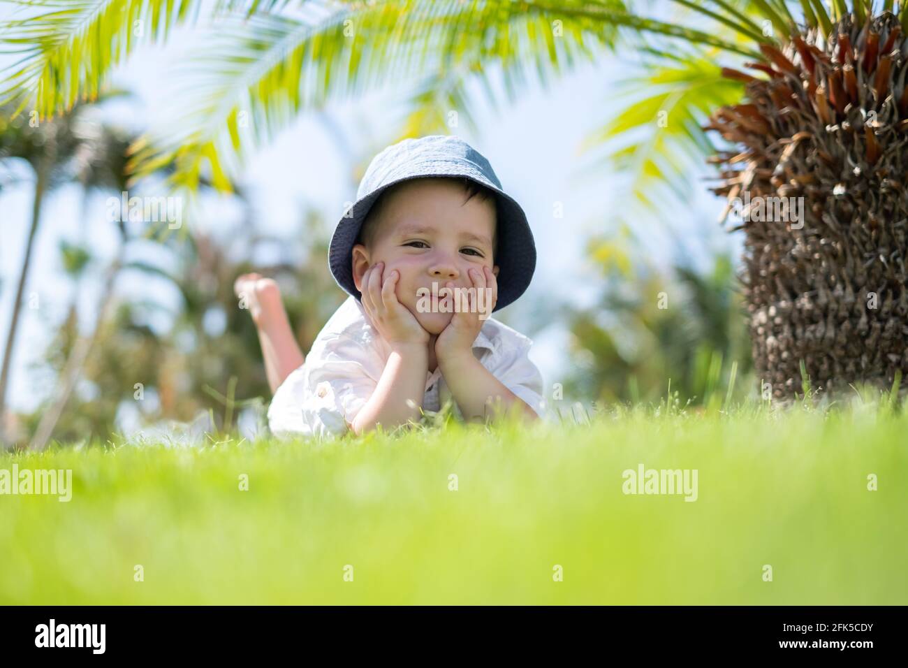 Un petit garçon heureux, allongé sur de l'herbe verte sous les palmiers Banque D'Images