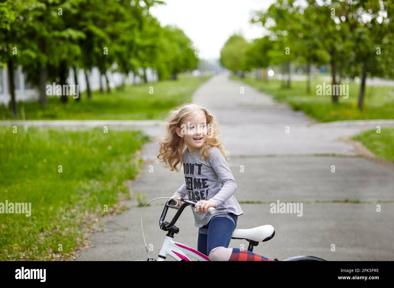 Une petite fille fait du vélo le jour d'été ensoleillé dans le parc de la ville. Les enfants apprennent à conduire un vélo sur une allée à l'extérieur Banque D'Images