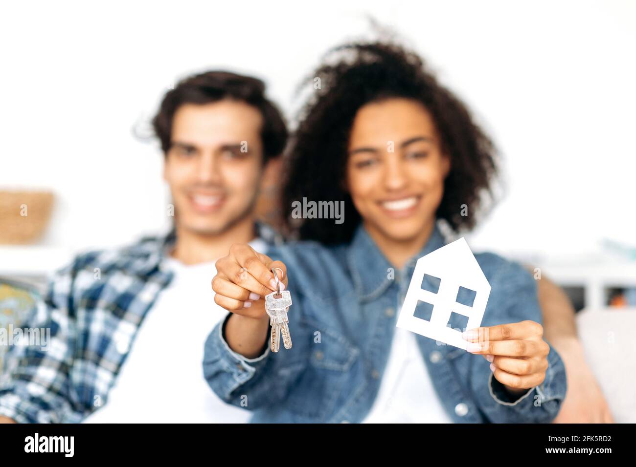 Joyeux heureux multiethnique jeune couple marié achète la propriété. Une fille afro-américaine floue tenant une maison en carton et des clés tout en étant assise sur le canapé avec son petit ami hispanique à la nouvelle maison Banque D'Images