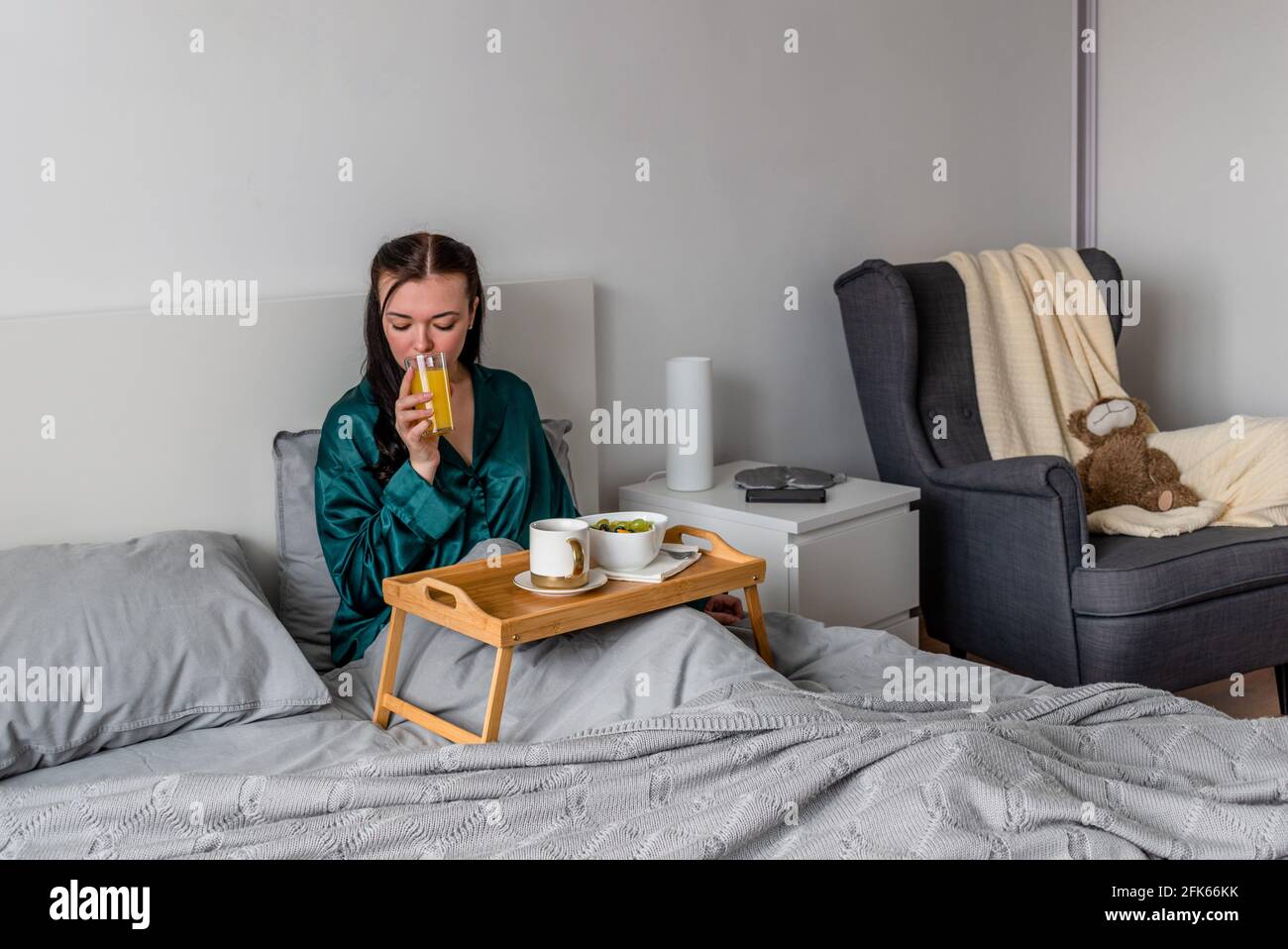 Une jeune femme en pyjama de soie mange son petit-déjeuner et boit du jus fraîchement pressé au lit. Concept bien-être. Banque D'Images