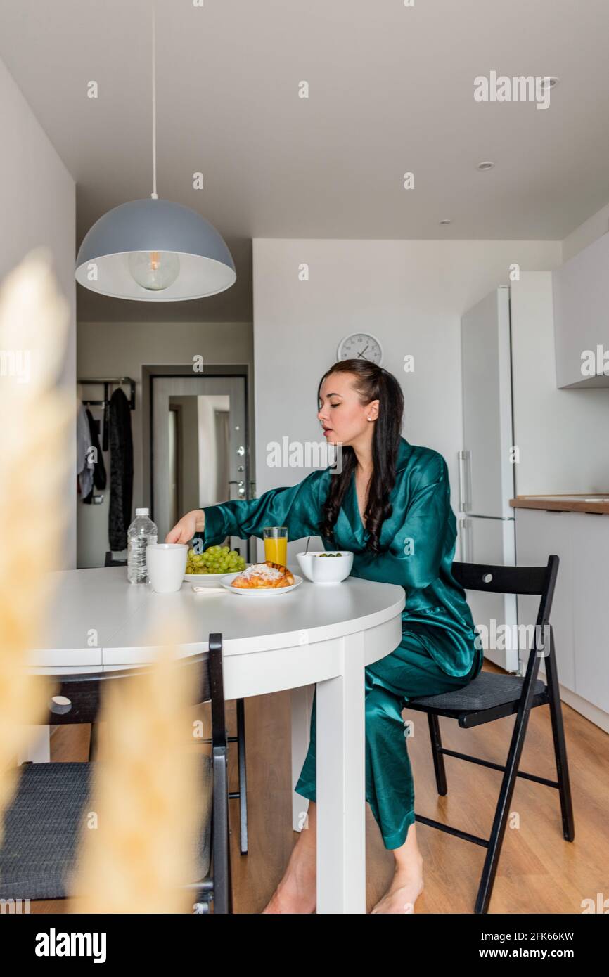 Jeune femme pyjama en soie a le petit déjeuner et boissons café dans la cuisine à la maison. Concept bien-être. Banque D'Images