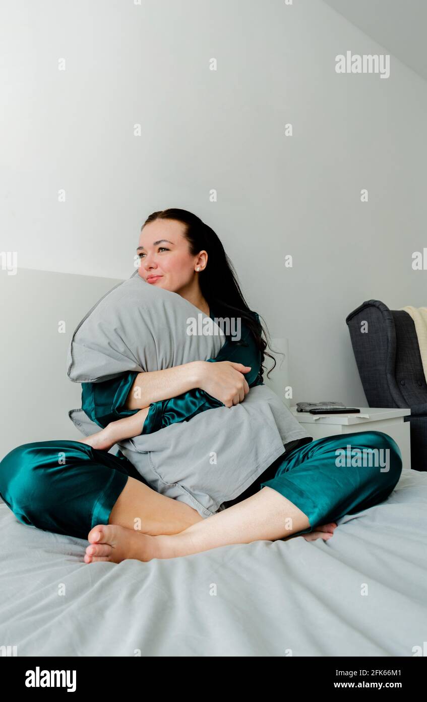 Une jeune femme en pyjama de soie est assise sur le lit et épouse un oreiller. Solitude à l'intérieur de la maison. Concept bien-être. Banque D'Images